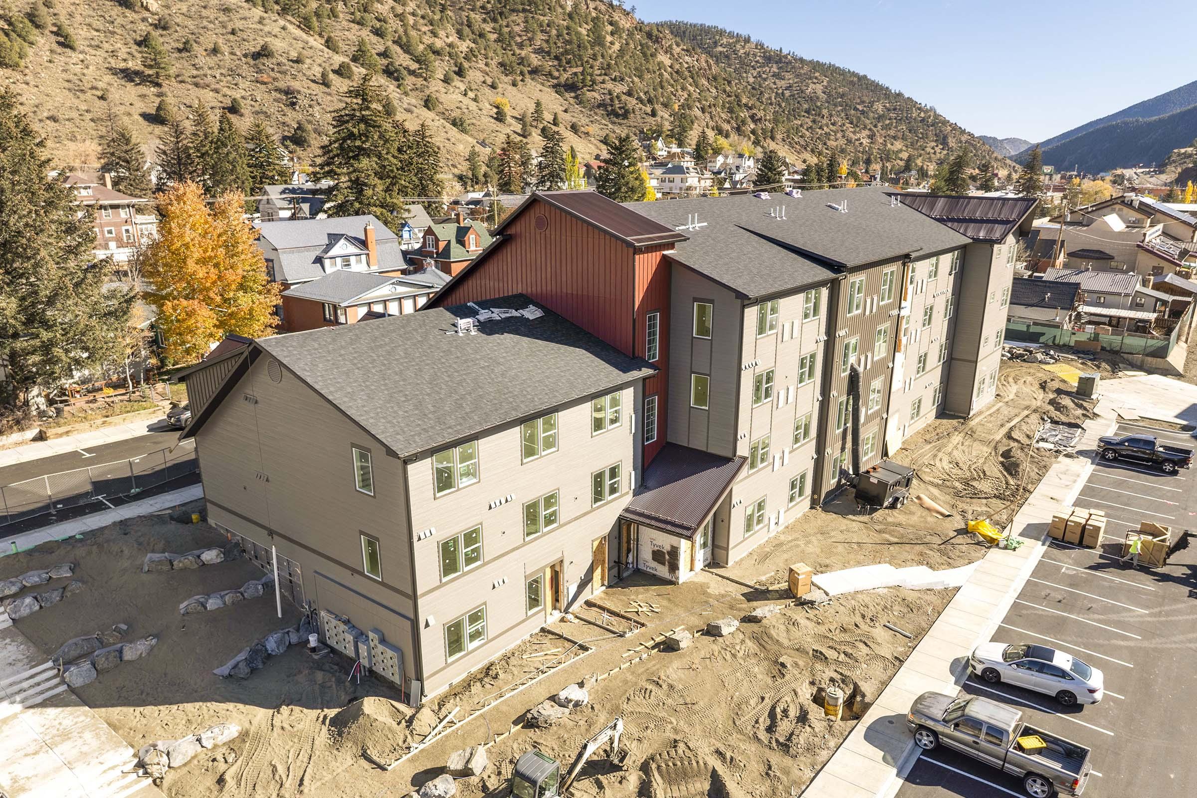 Aerial view of a large multi-story building under construction, surrounded by raw earth and construction materials. The building features a mix of brown and gray siding with a red accent on one side. In the background, a hilly landscape and nearby houses are visible, with autumn foliage highlighting the scene.