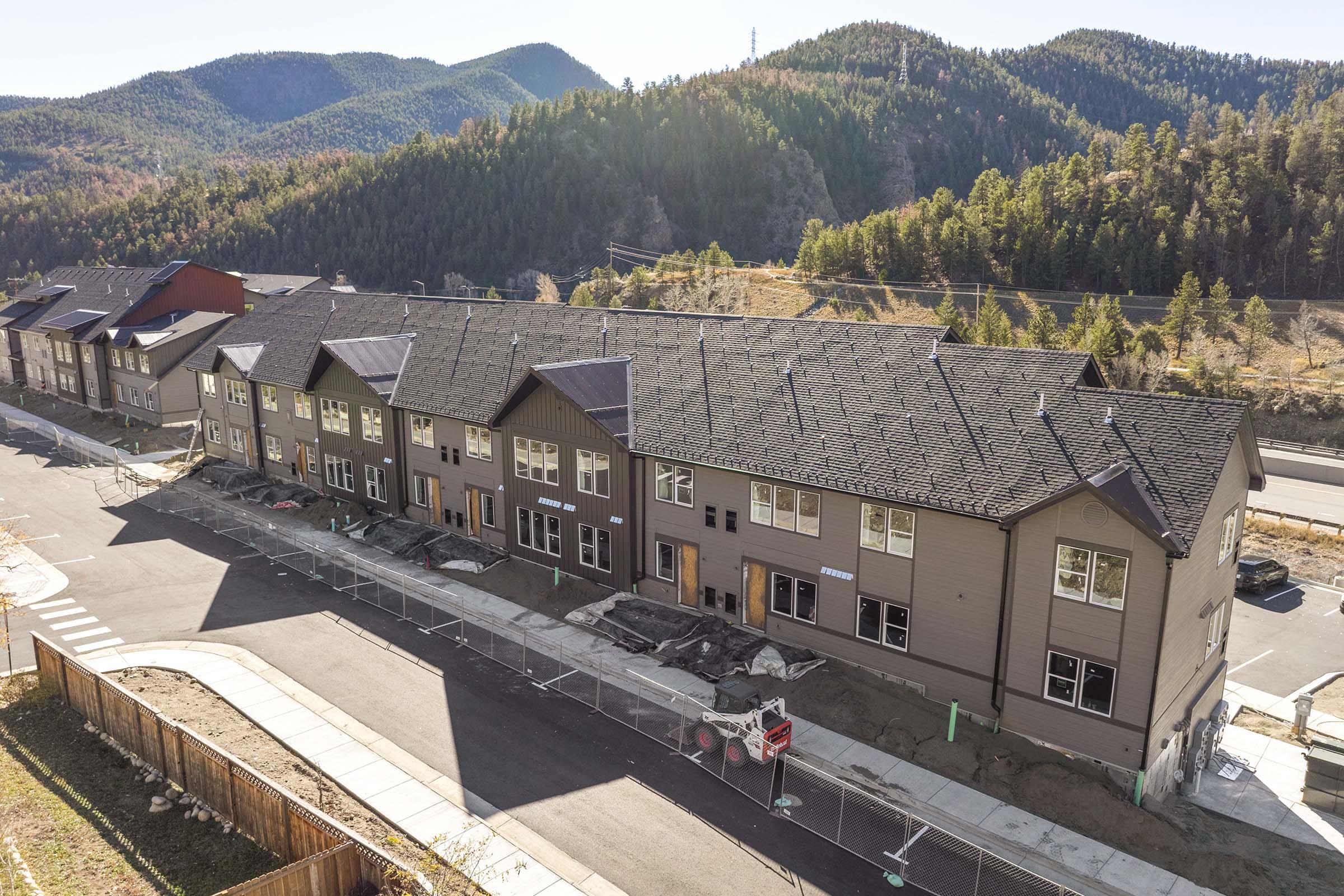 Row of newly constructed townhouses in a mountainous area, with unfinished landscaping and construction materials visible. The buildings have multiple windows and a modern design, surrounded by trees and hills in the background. A road runs alongside the development.