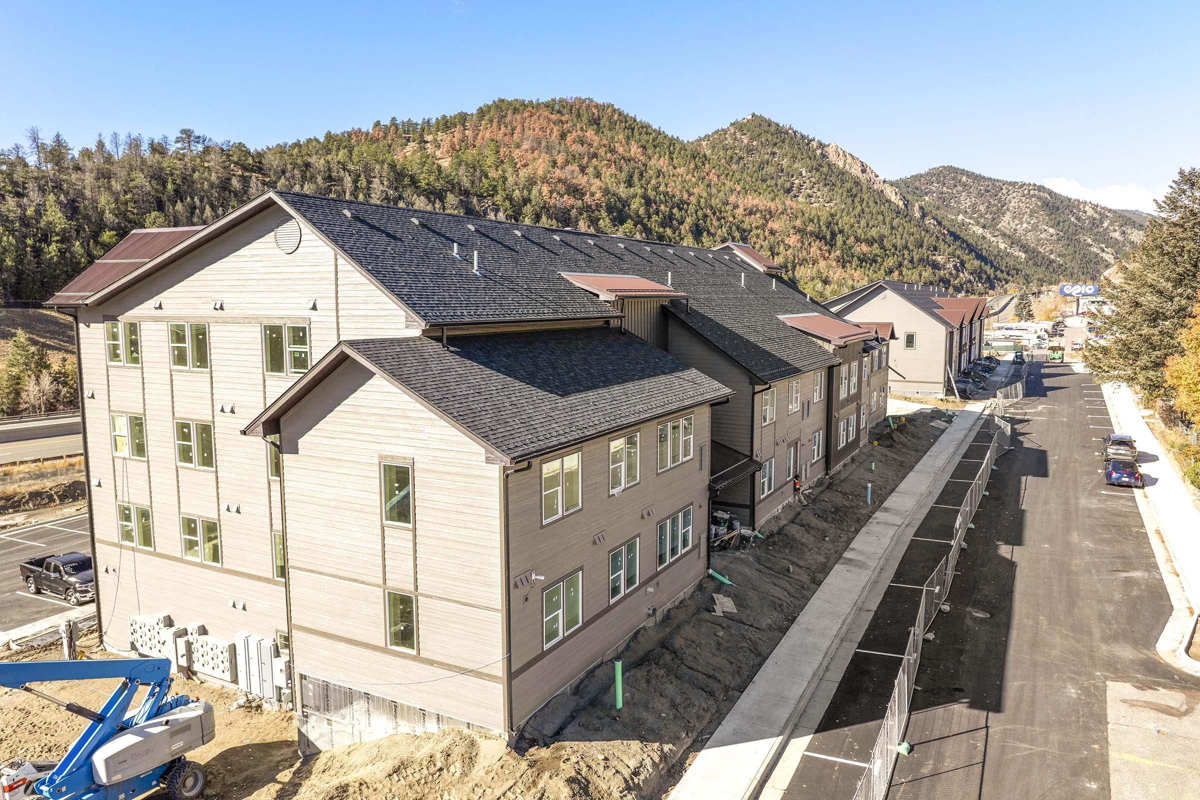 A side view of a multi-story residential building under construction, featuring a light-colored exterior and green windows. The structure is situated near a paved road with parked vehicles, surrounded by trees and mountains in the background, showcasing an autumn landscape with colorful foliage.