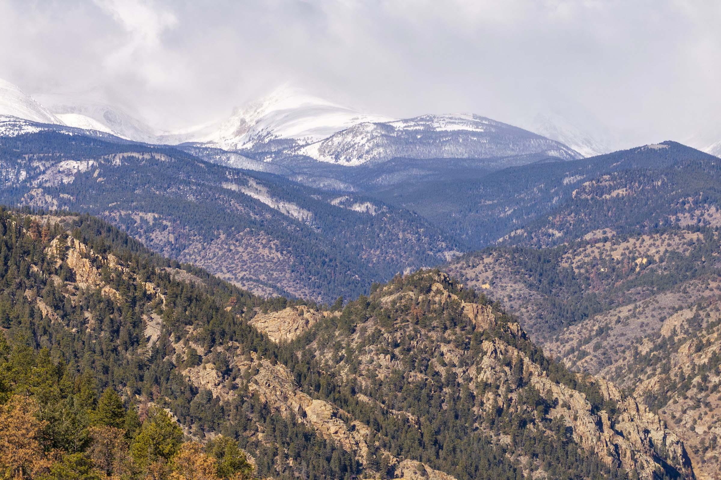 A panoramic view of rugged mountain ranges with snow-capped peaks in the background. The lower slopes are covered in dense evergreen forests, while the terrain has rocky outcrops and varying shades of green and brown, suggesting a mix of vegetation and earth. The sky is overcast, adding a dramatic ambiance to the scene.
