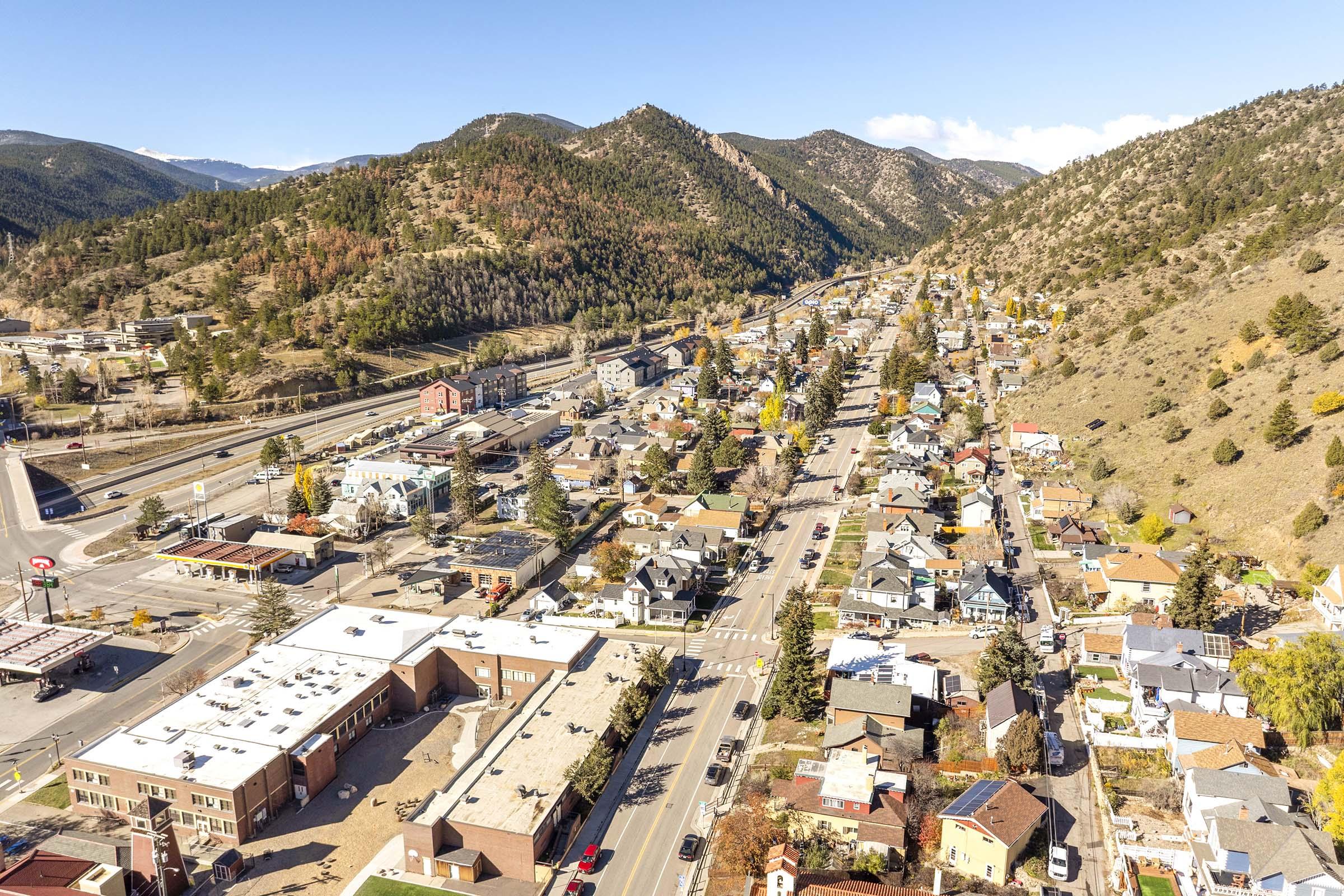 Aerial view of a small town nestled between mountains, showcasing a mix of residential houses and commercial buildings along a main road. The scene includes green hills in the background, a clear blue sky, and trees displaying autumn colors, creating a vibrant landscape.