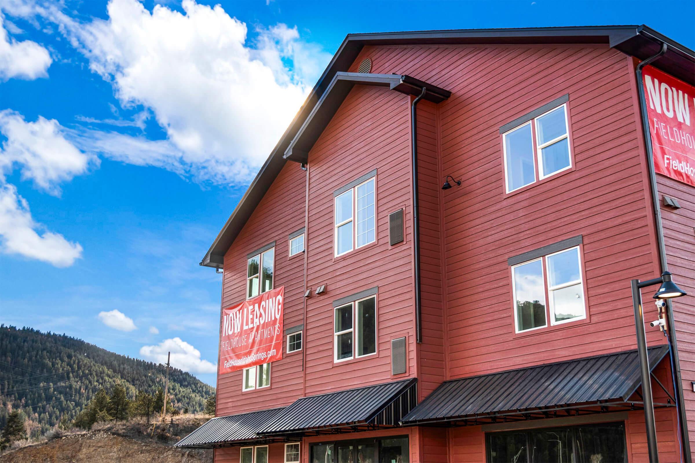 A modern red building with large windows, showcasing a "NOW LEASING" banner. The structure is set against a backdrop of trees and a blue sky with white clouds. The design features a peaked roof and a metal awning, emphasizing a contemporary architectural style.