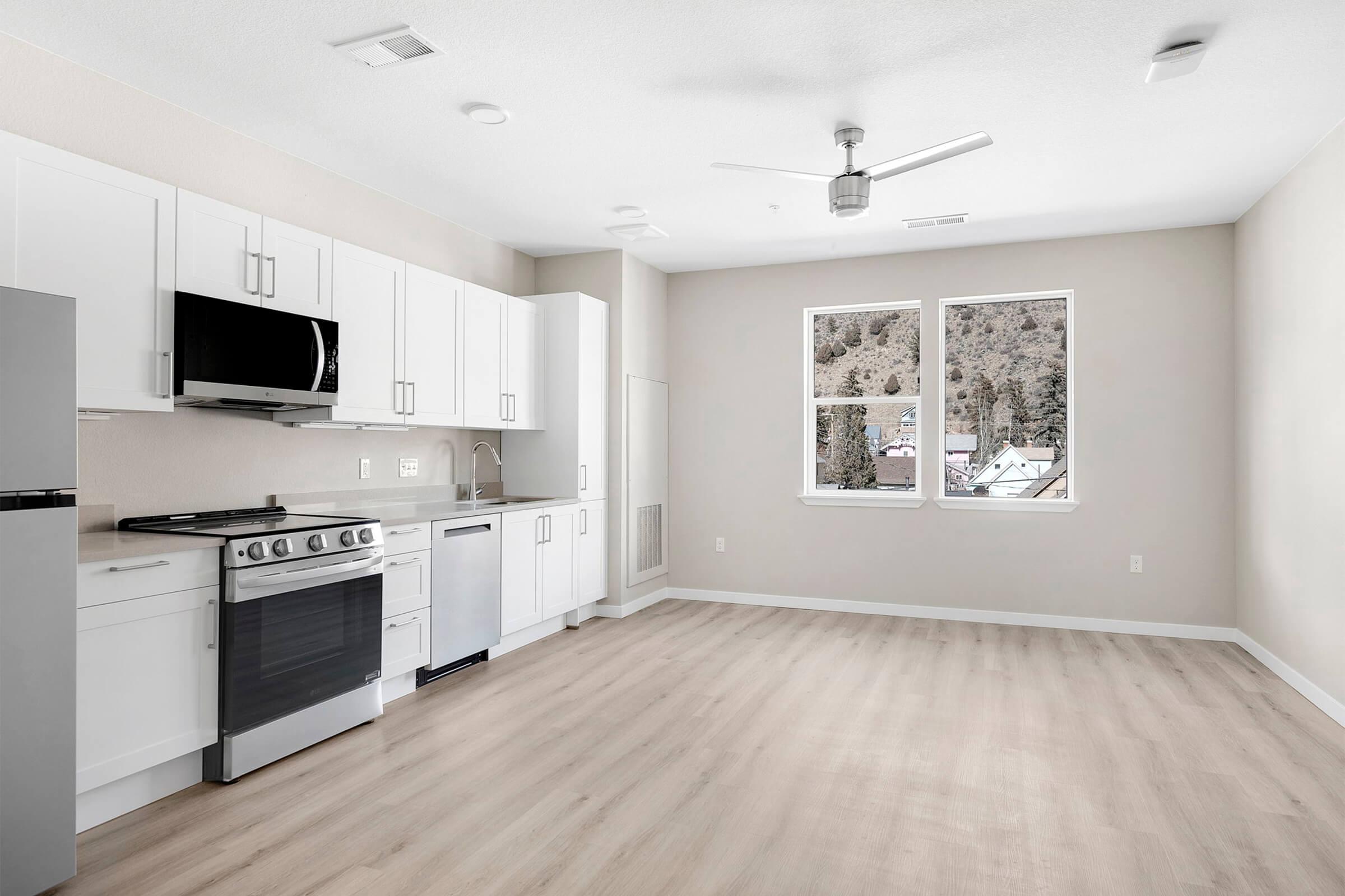 A modern kitchen and living space with white cabinetry, stainless steel appliances including a gas stove and refrigerator, and large windows providing natural light. The floor is light-colored wood, and the walls are painted in a neutral tone, creating a bright and airy atmosphere.
