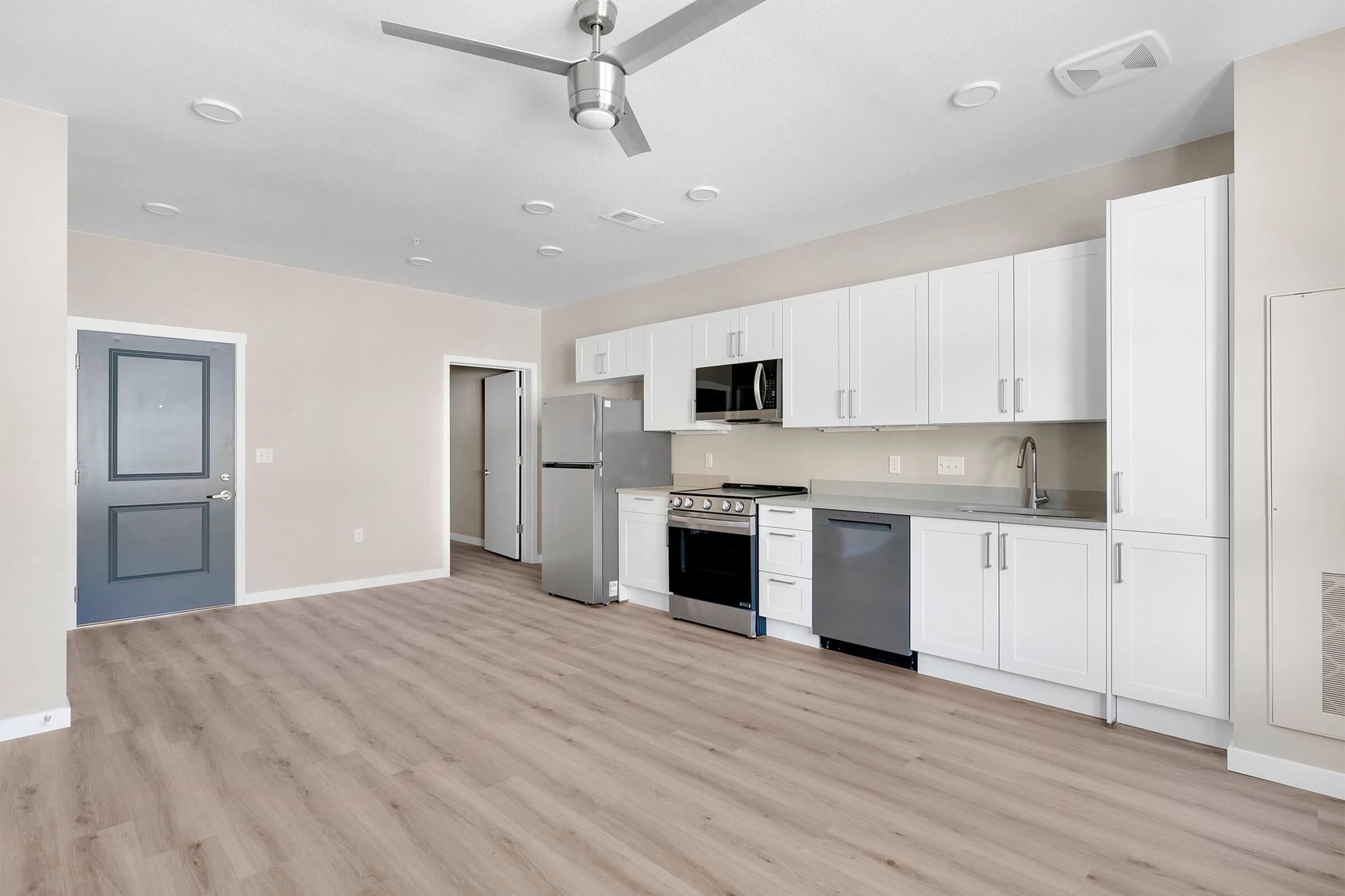 Modern kitchen with white cabinetry, stainless steel appliances including a refrigerator, stove, and microwave. The flooring is light wood, and natural light comes from a doorway leading to another room. A ceiling fan is installed, adding to the contemporary design of the space.