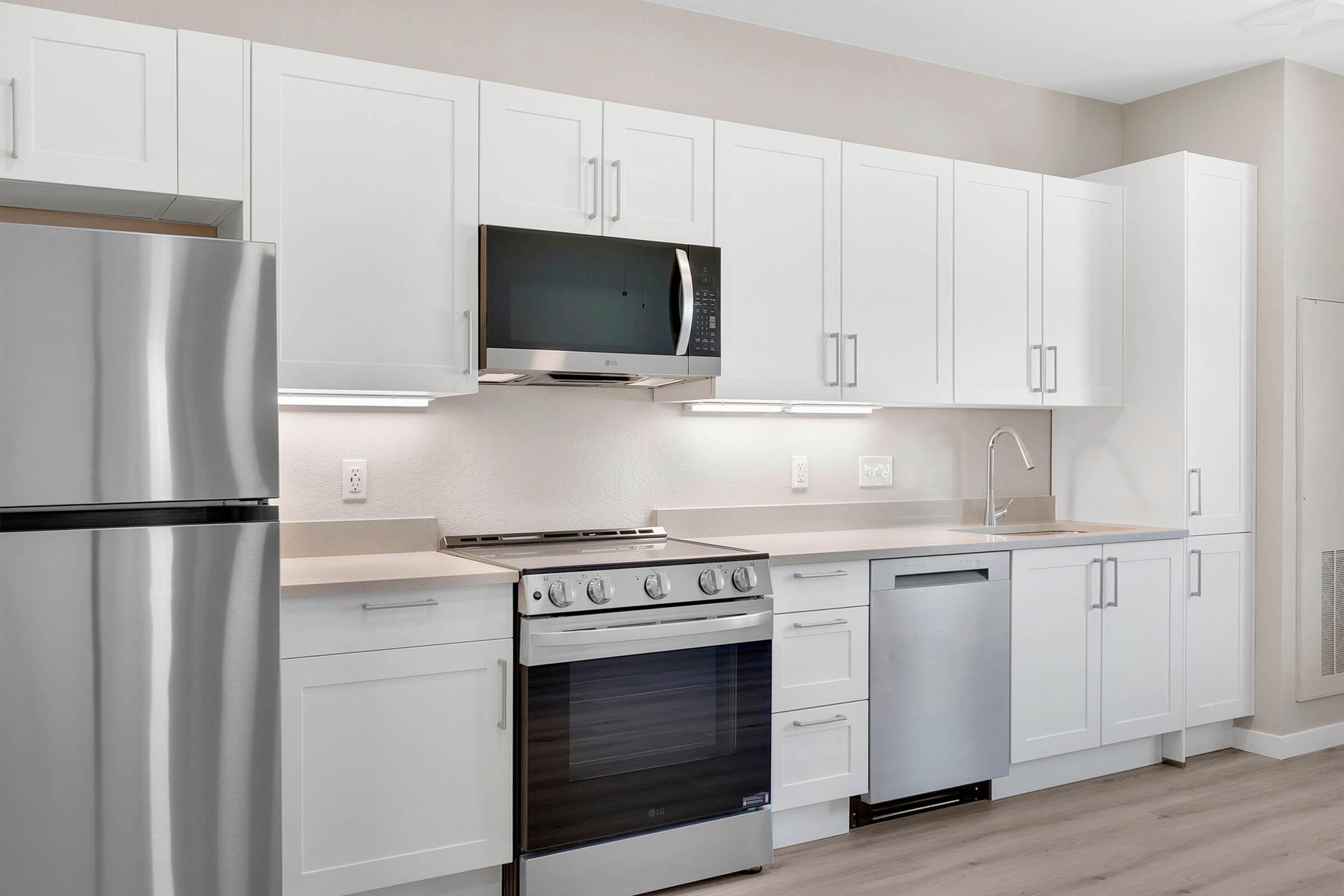 A modern kitchen featuring white cabinets, stainless steel appliances including a refrigerator and oven, a built-in microwave, and a sleek countertop. The space is well-lit with warm lighting, creating a clean and inviting atmosphere. Light-colored flooring complements the overall design.