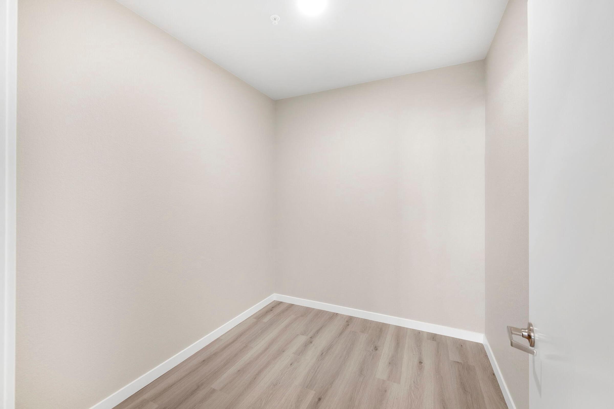 Empty room with light beige walls, wooden flooring, and a single light fixture in the ceiling. An open white door is visible on the right side, leading into the room. The space appears clean and minimalistic, suitable for various uses.