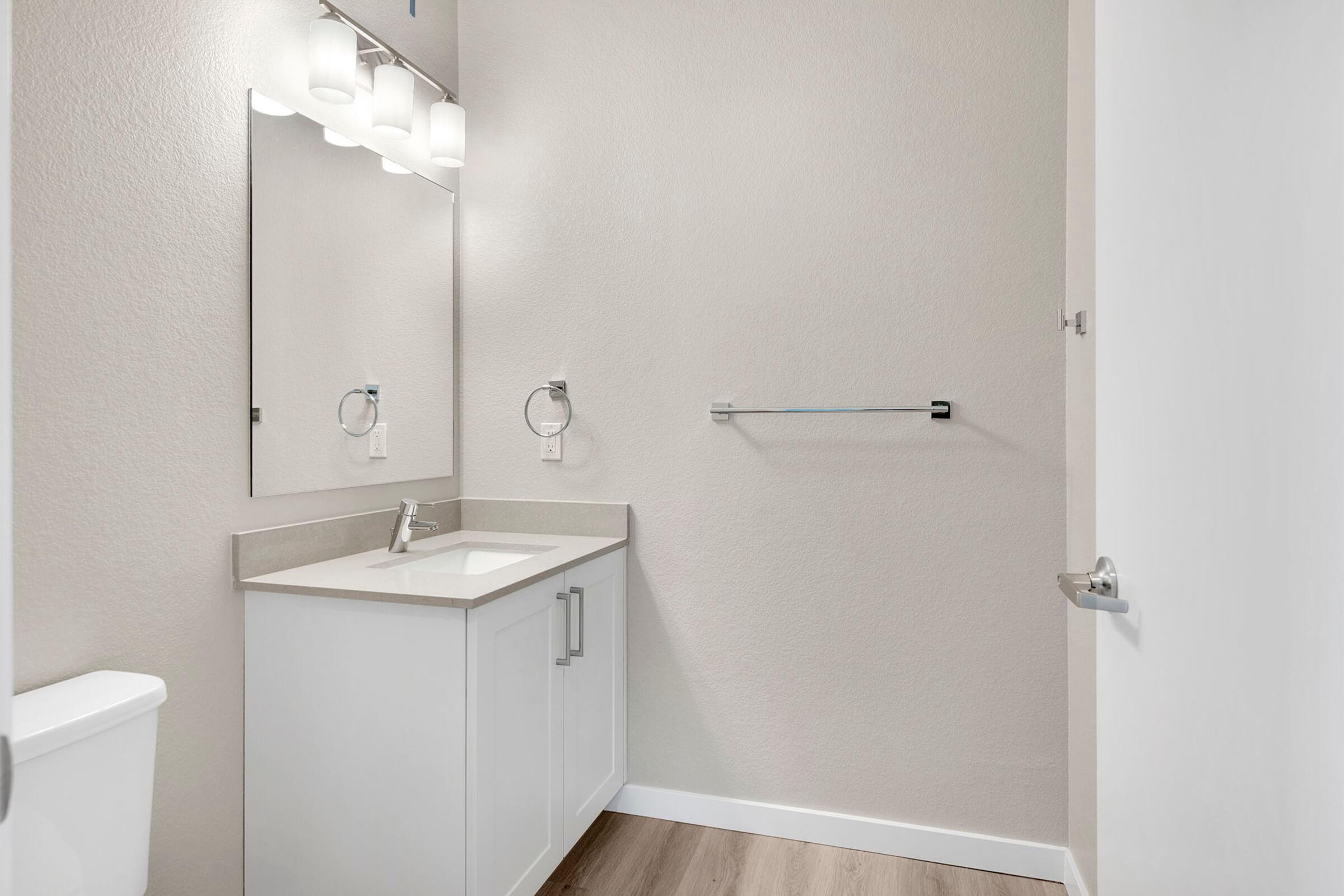 A clean, modern bathroom featuring a white vanity with a sink, a large wall mirror, and three light fixtures above. The walls are painted in a light neutral color, and there is a towel bar installed. The floor is made of wooden planks, and a white toilet is visible on the left side.