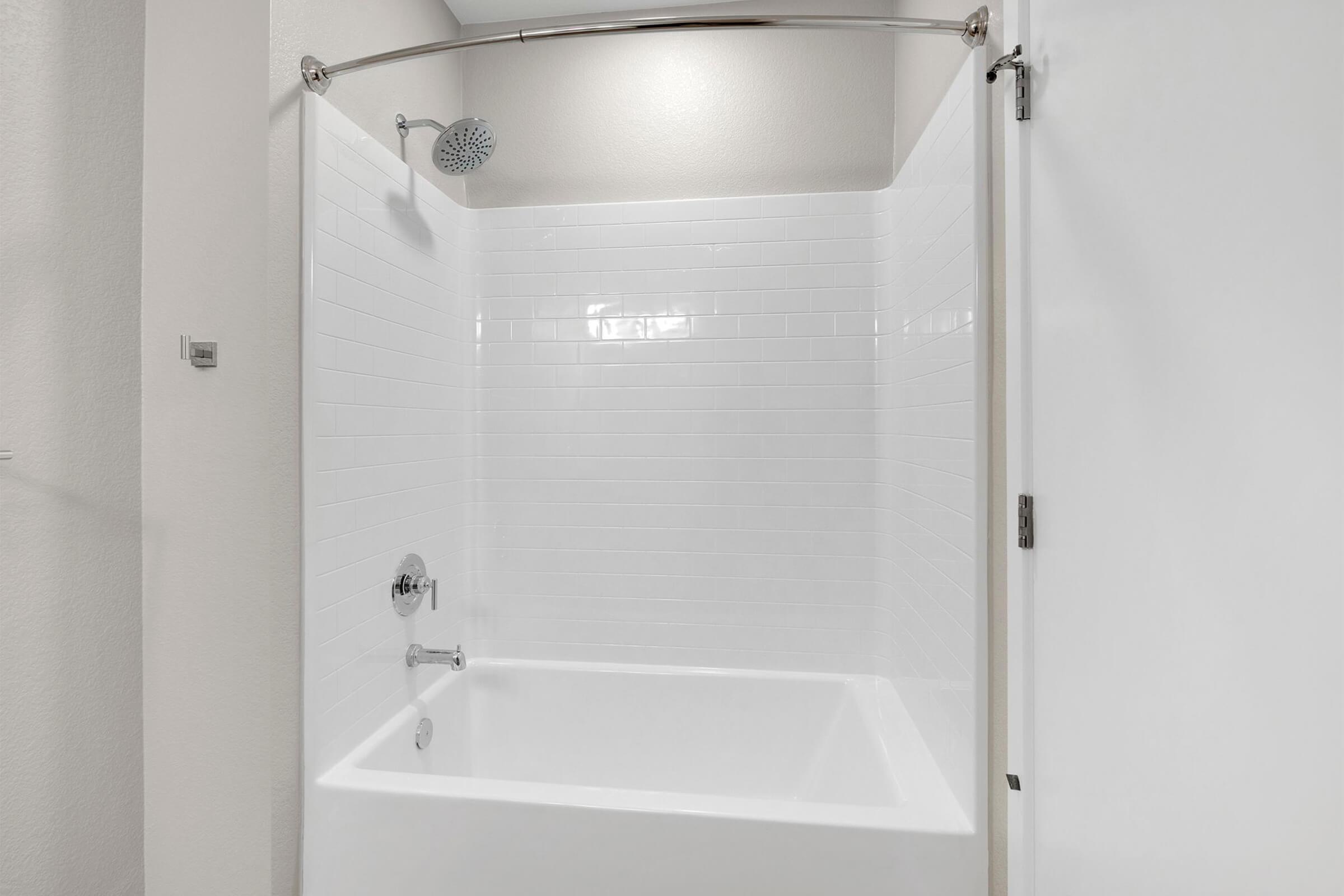 A clean, modern bathroom featuring a white tiled shower area with a curved shower rod and a wall-mounted showerhead. The space is well-lit and minimalist, with a plain white tub and a closed door nearby, creating a tidy and inviting atmosphere.