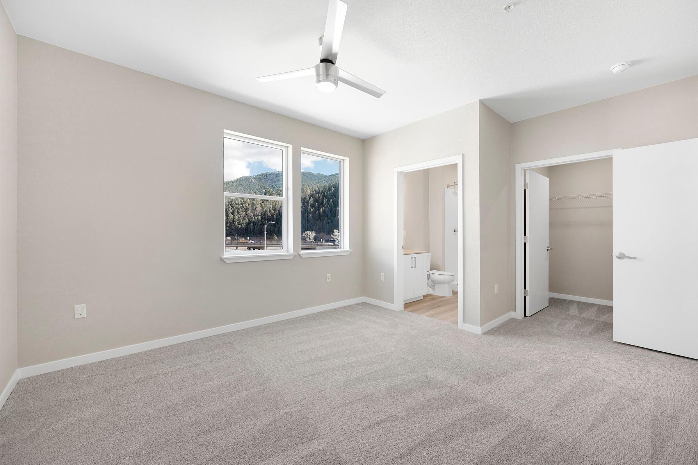 A spacious, empty room with light-colored walls and carpet. It features large windows allowing natural light, a ceiling fan, and an adjacent bathroom area that includes a toilet and a closet space. The view outside shows trees and mountains in the distance.