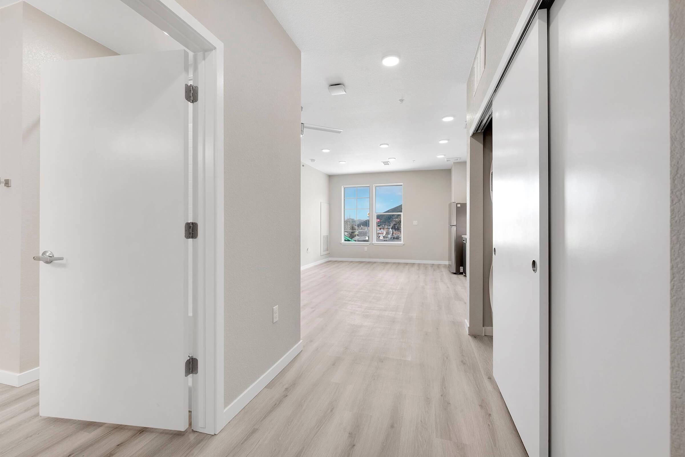 A view of a modern, spacious interior hallway leading to a well-lit living area. The walls are painted in neutral tones, and there are large windows allowing natural light. Closet doors are visible, and the flooring is light wood. A door on the left leads to another room or area.