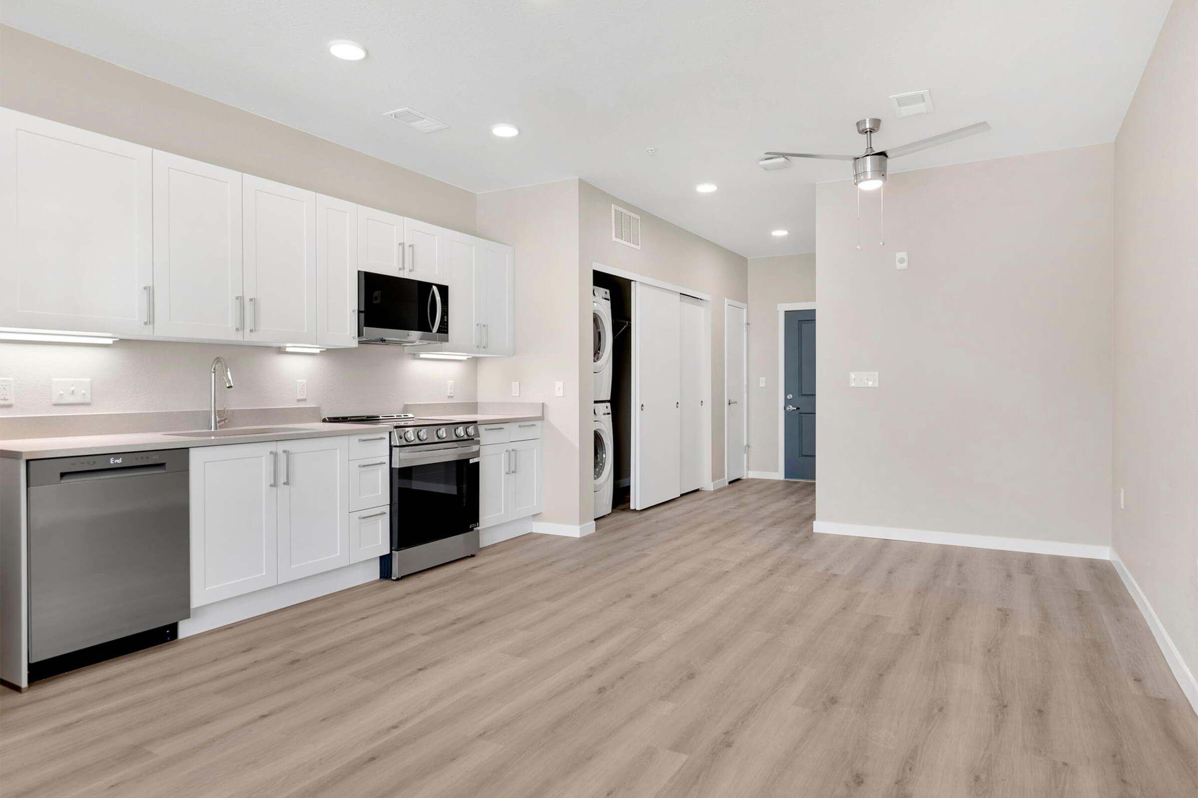Modern kitchen featuring white cabinetry, stainless steel appliances, and a light-colored countertop. The open layout includes a living area with light wood flooring and a ceiling fan. Nearby, there are laundry appliances in adjacent closet spaces, contributing to a clean and spacious design.