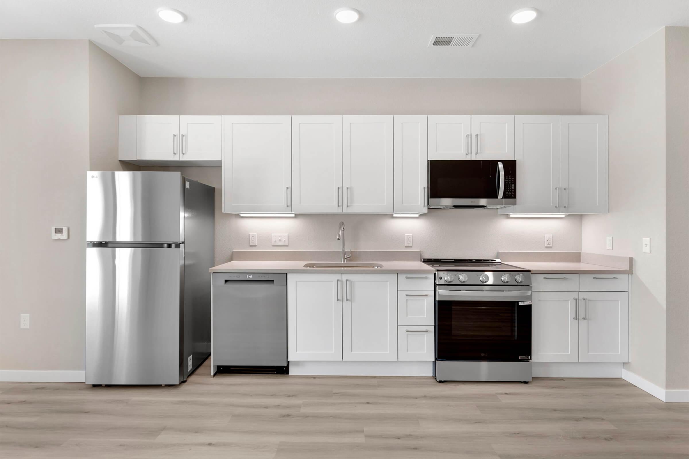 Modern kitchen featuring stainless steel appliances including a refrigerator, microwave, and oven. The cabinets are white with a sleek design, complemented by a light-colored countertop. The space has neutral wall tones and bright overhead lights, creating a clean and contemporary atmosphere.