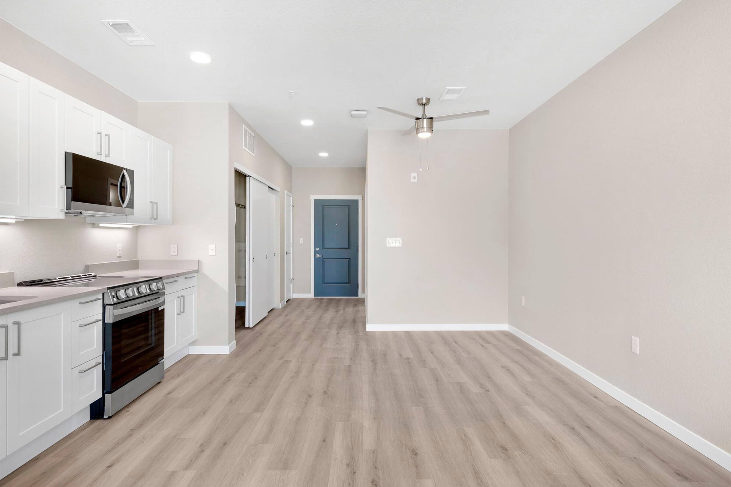 A modern kitchen area with stainless steel appliances, including a stove and microwave, alongside white cabinetry. Light wood flooring runs throughout the open space, which connects to a living area with a ceiling fan and a blue door leading to another room. Walls are painted in neutral tones.