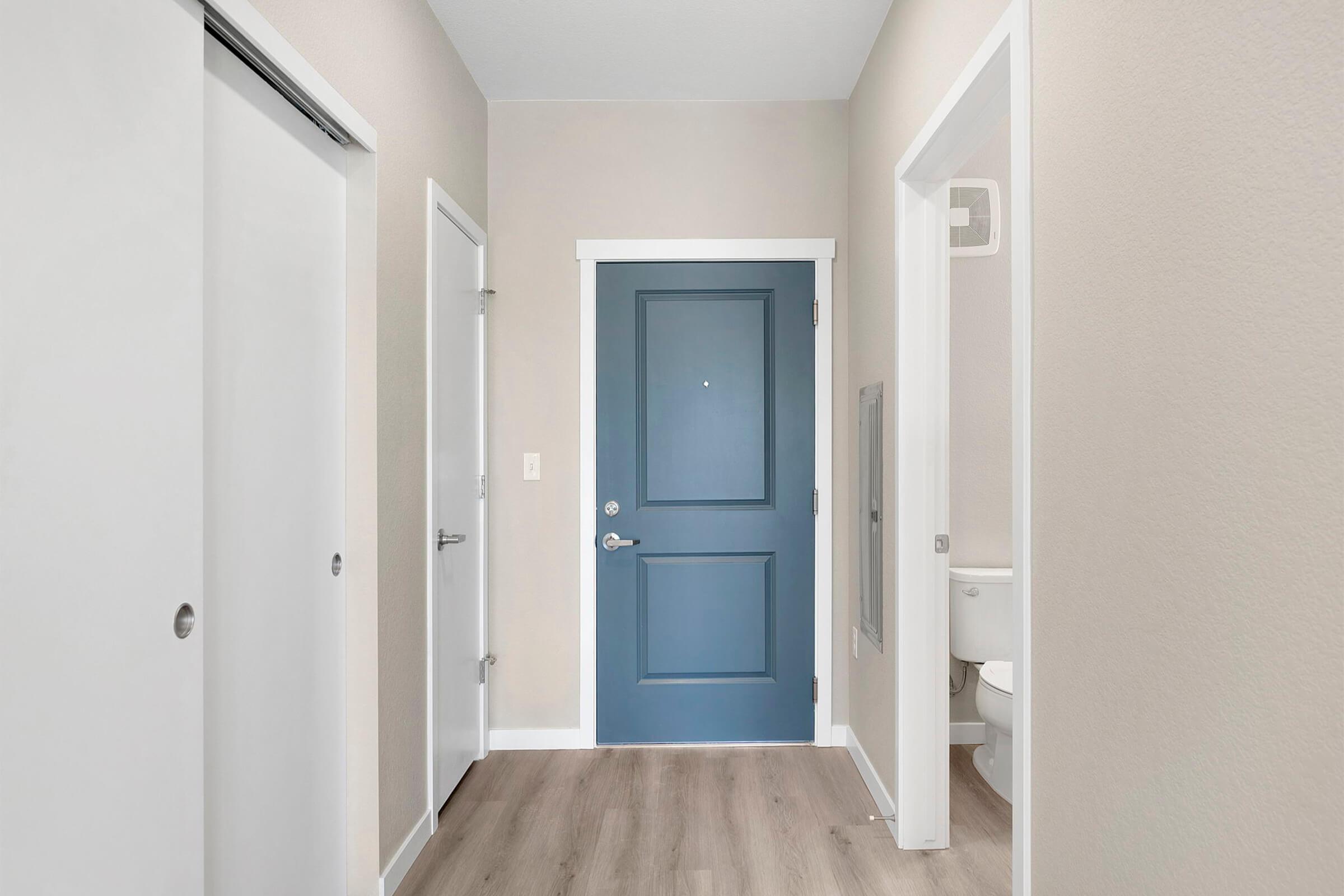 A well-lit hallway with light beige walls and hardwood flooring. The main feature is a dark blue front door at the end of the hallway. There are two white doors on the left, one with a circular handle and another with a vertical handle. A bathroom door is partially visible on the right.