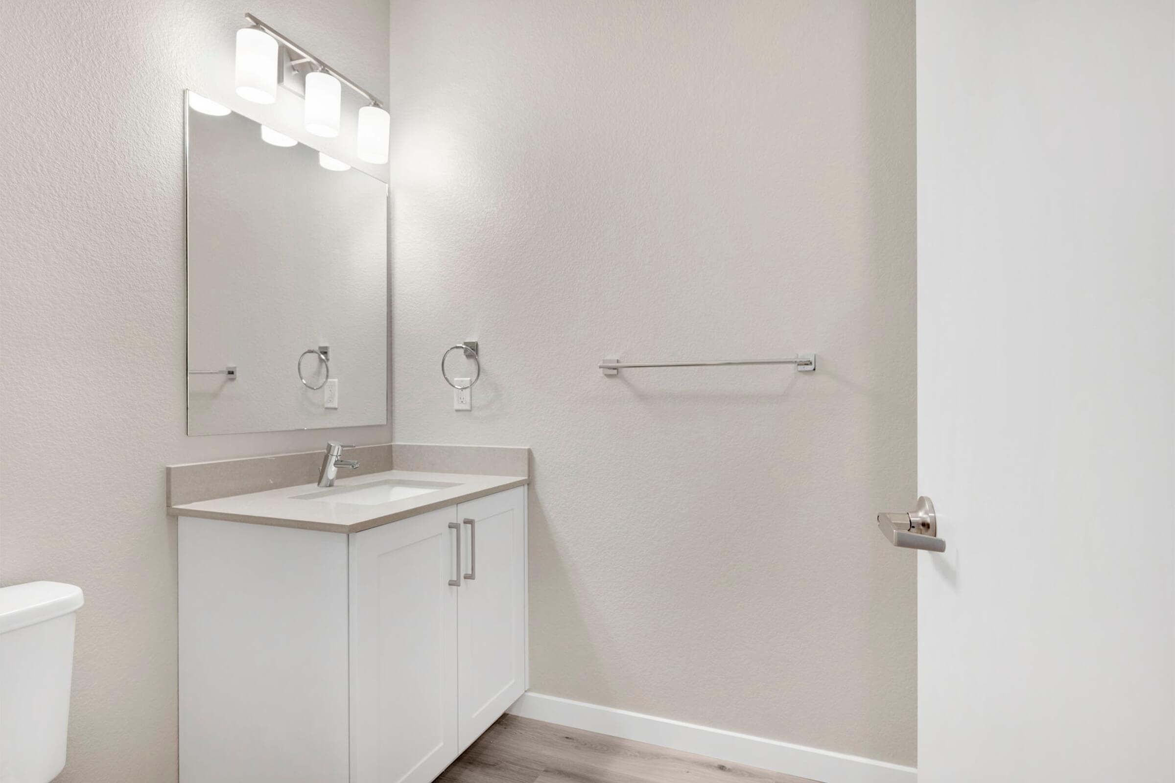 Bright and modern bathroom featuring a single sink vanity with a large mirror above, stylish light fixtures, and a wall-mounted towel bar. The walls are painted in a neutral color, and the flooring is a light wood finish. A white toilet is visible on the left, with a sleek door on the right.