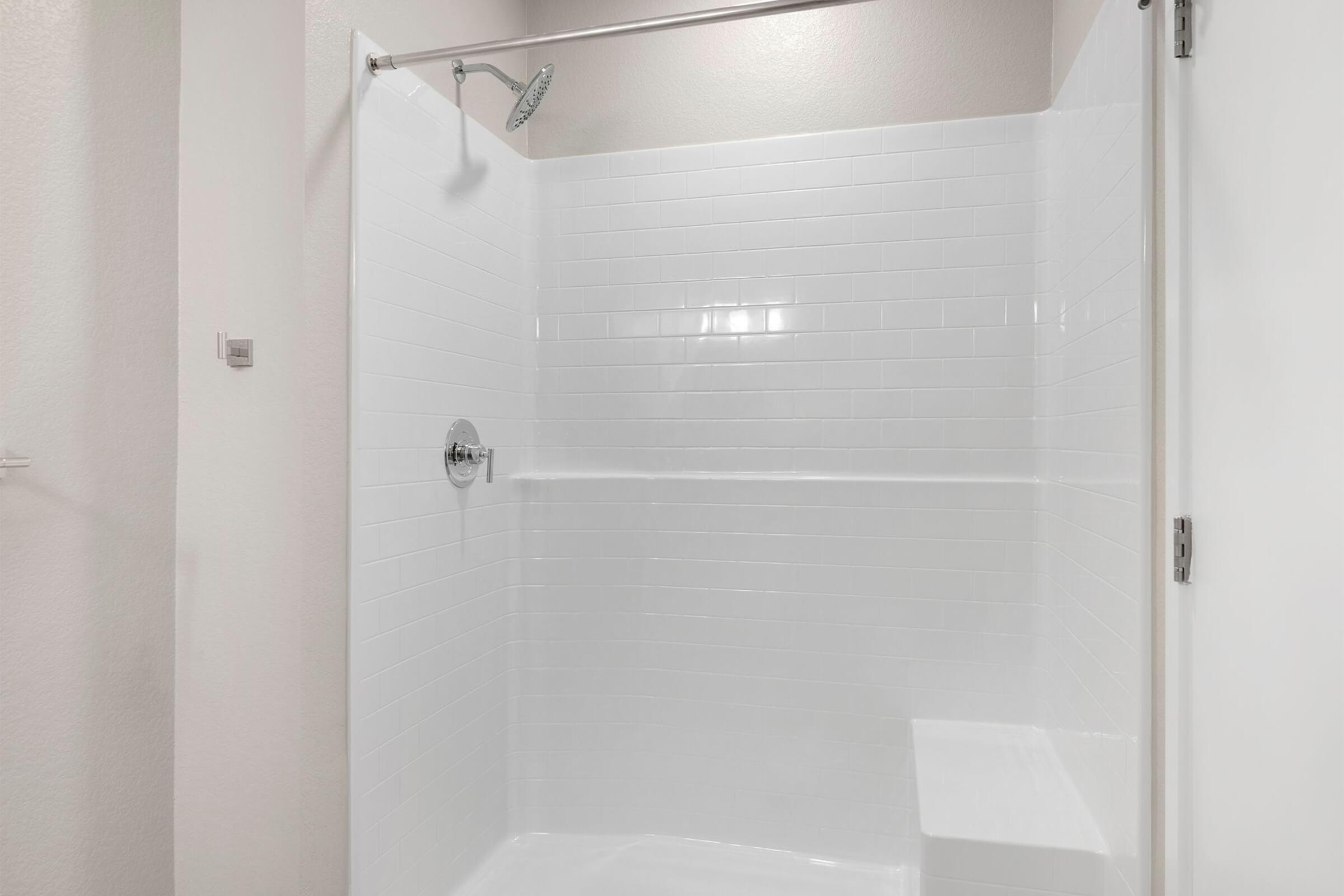 A clean and modern shower area featuring white tiled walls, a wall-mounted showerhead, and a small built-in step. The space is well-lit with a bright, neutral color scheme, creating a spacious and inviting atmosphere.