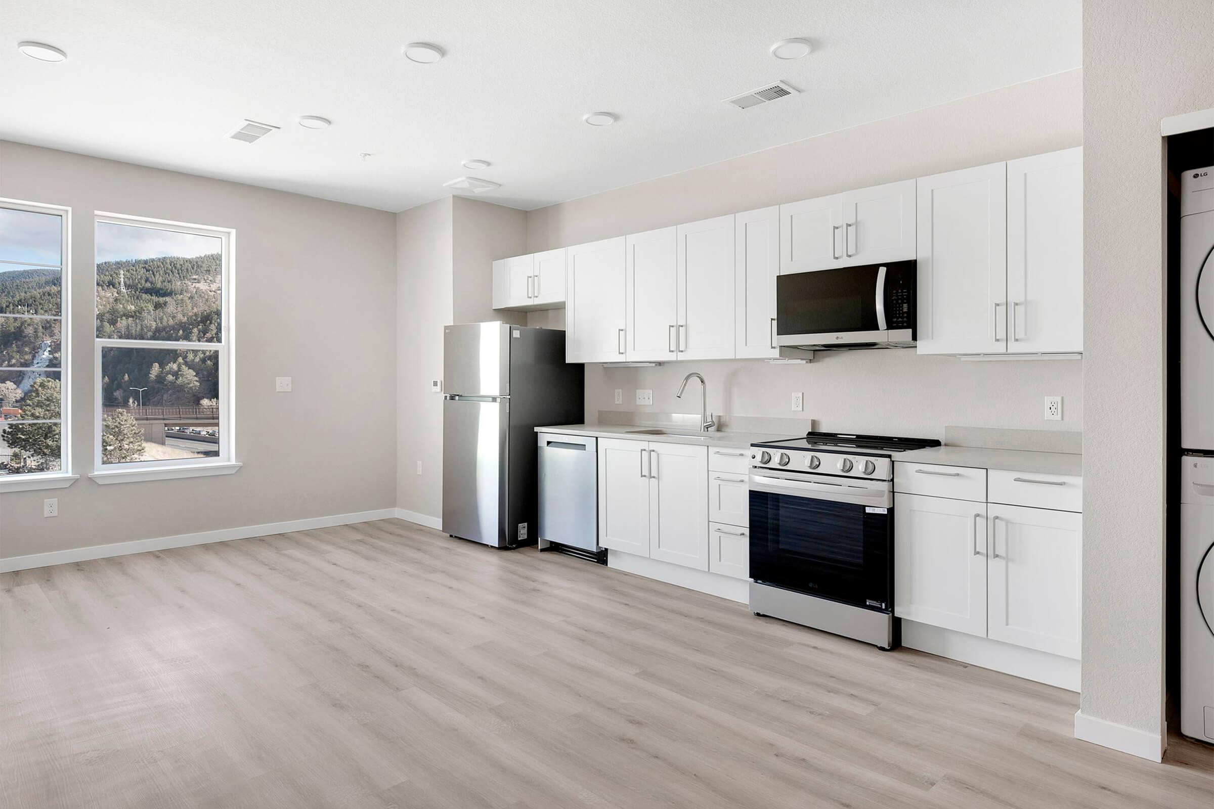 Modern kitchen in a spacious apartment featuring white cabinets, stainless steel appliances, a refrigerator, a dishwasher, an oven, and a microwave. Large windows provide natural light and a view of greenery outside. The flooring is light-colored wood.