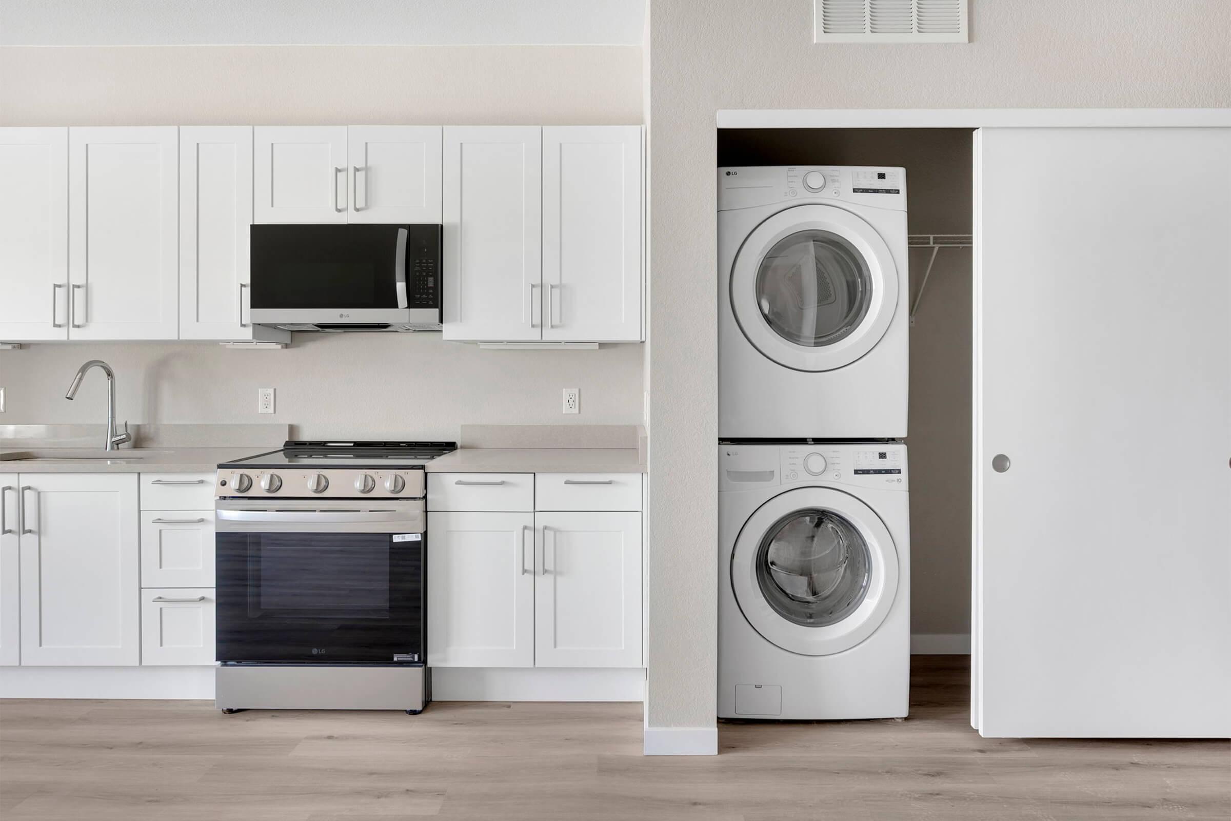 Modern kitchen with white cabinetry, stainless steel microwave, and a gas stove. Adjacent is a compact laundry area featuring stacked white washer and dryer. The space has light-colored flooring and a neutral wall, creating a clean, bright atmosphere.