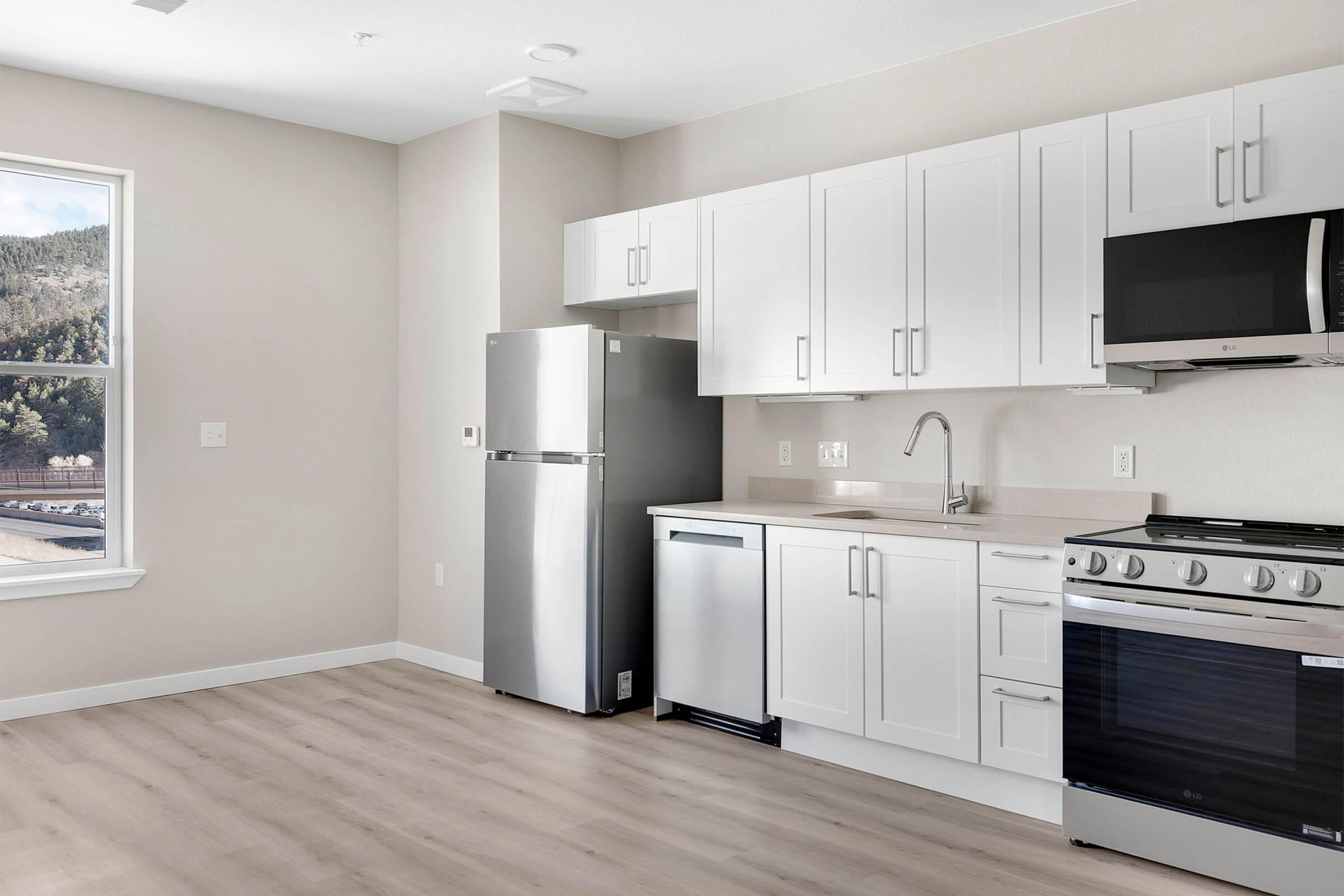 Modern kitchen with white cabinetry, stainless steel refrigerator, dishwasher, and oven. A sink is installed under a window that lets in natural light, highlighting the light-colored walls and flooring. The space is clean and minimally decorated, creating a fresh, contemporary look.