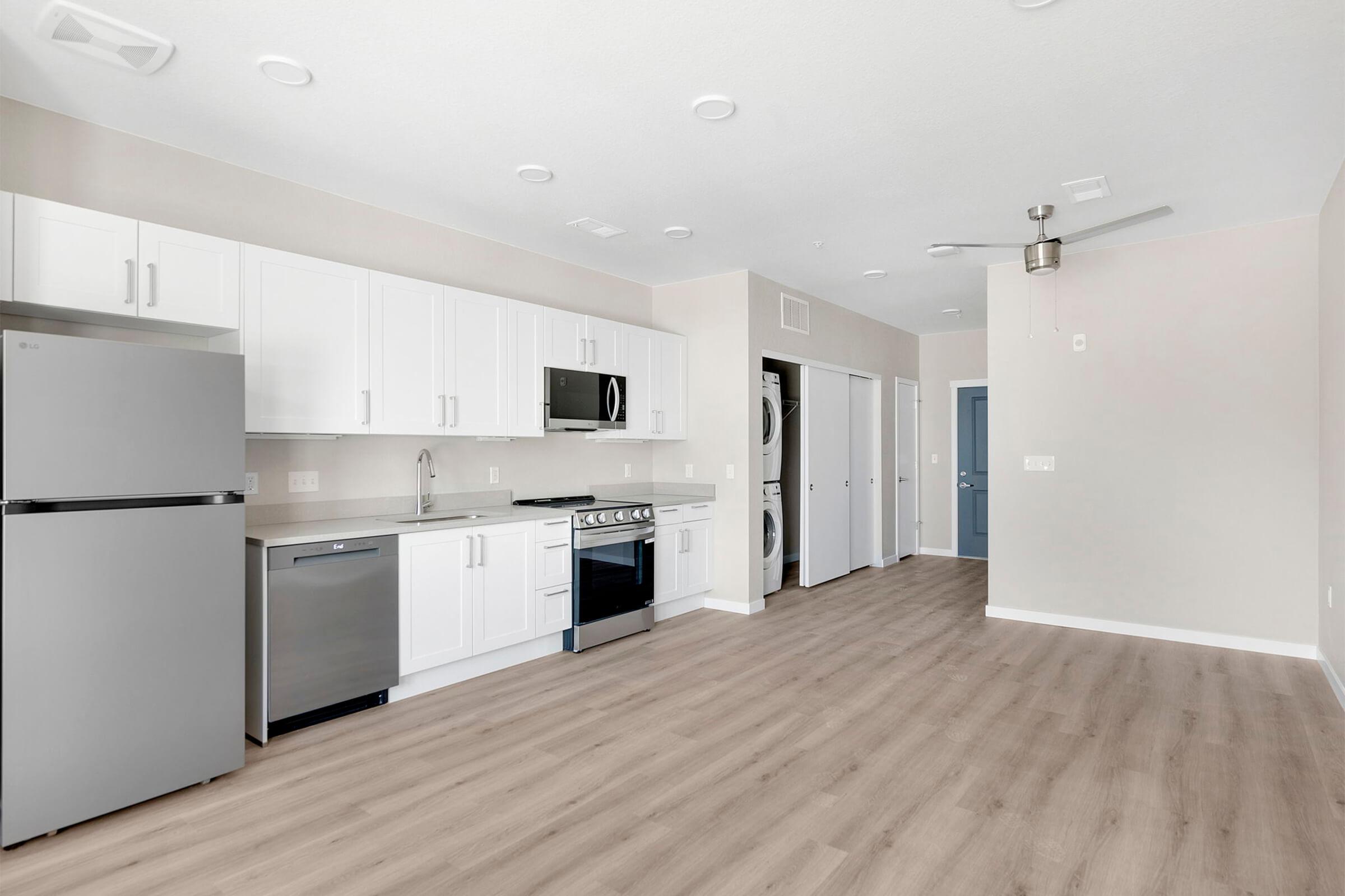 Modern kitchen and living area featuring stainless steel appliances, including a refrigerator, oven, and microwave. White cabinetry contrasts with light-colored walls and wood-style flooring. A ceiling fan is installed, and there are doors leading to other rooms in the background.