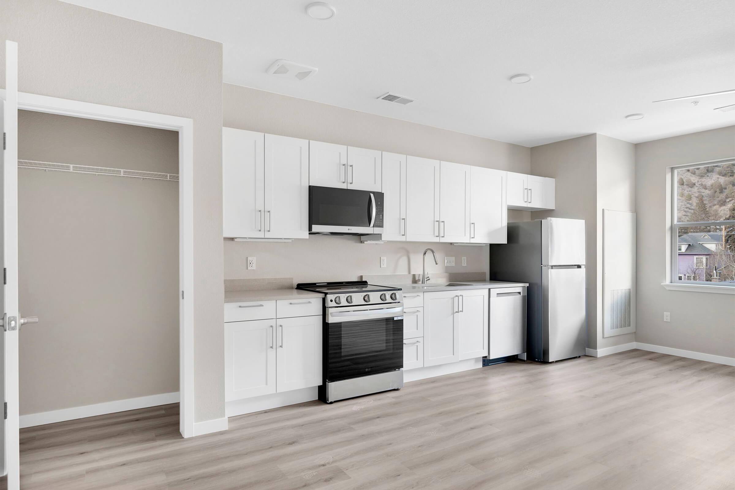 Modern kitchen in a bright, open space featuring white cabinetry, stainless steel appliances, and a large window. The layout includes a stove, microwave, refrigerator, and a sink, with light-colored flooring and neutral wall tones, creating a clean and inviting atmosphere.