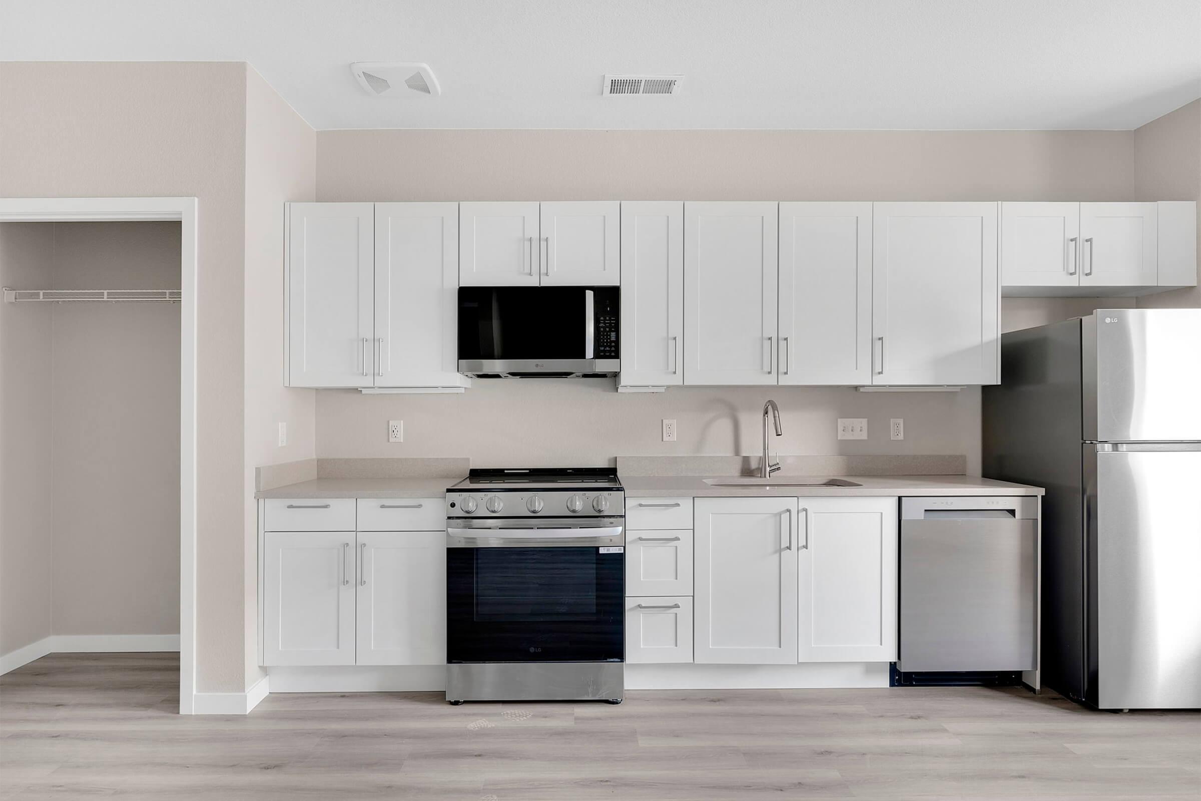 Modern kitchen featuring white cabinets, a stainless steel refrigerator, an oven with a cooktop, a microwave above the stove, and a dishwasher. Light-colored walls and flooring create a bright, airy atmosphere. A closet space is visible in the corner.