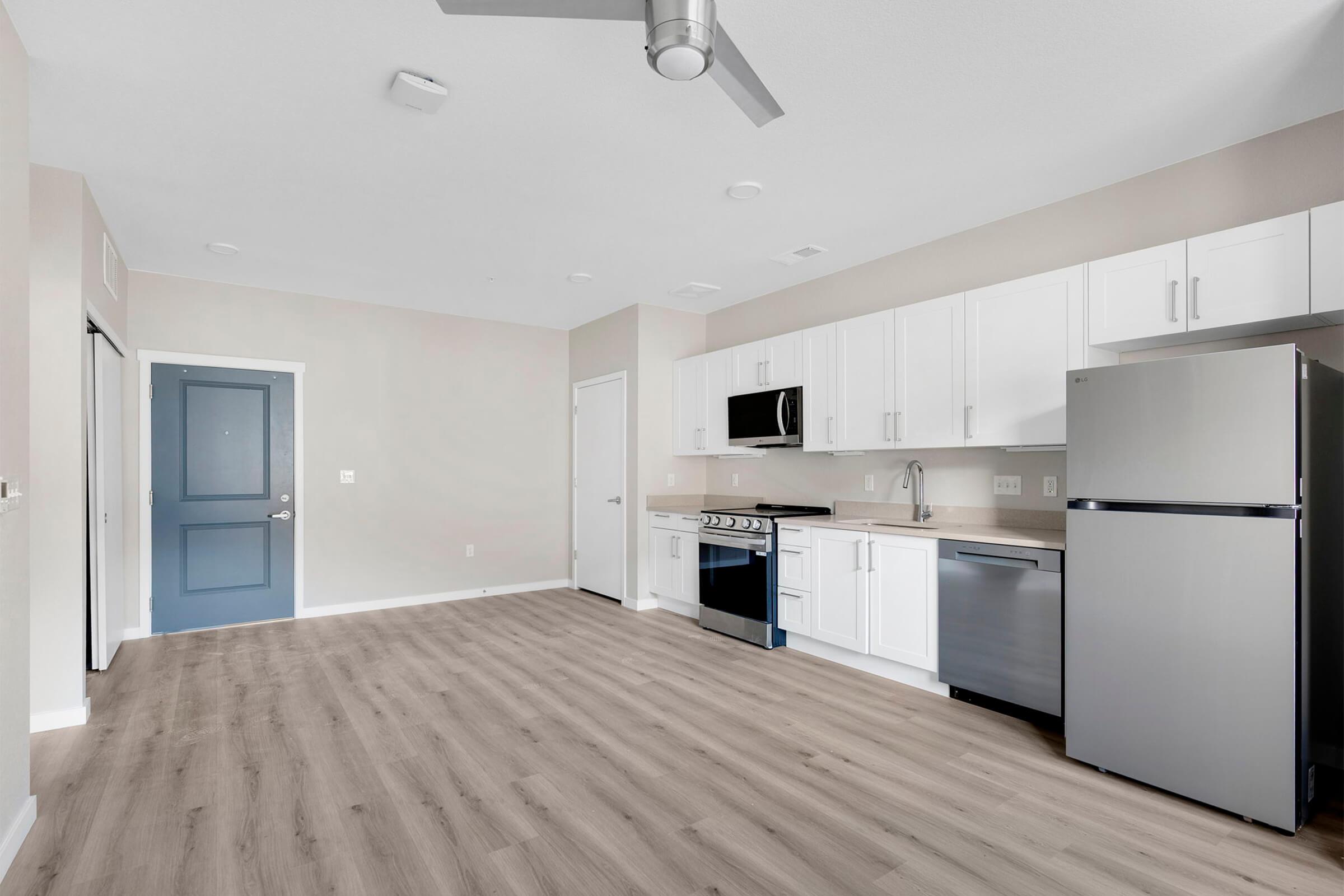 Modern kitchen with white cabinetry, stainless steel appliances, and a spacious layout. Includes a refrigerator, microwave, and stove, with wooden flooring and a blue front door visible. The walls are painted in a light neutral color, creating a bright and inviting atmosphere.