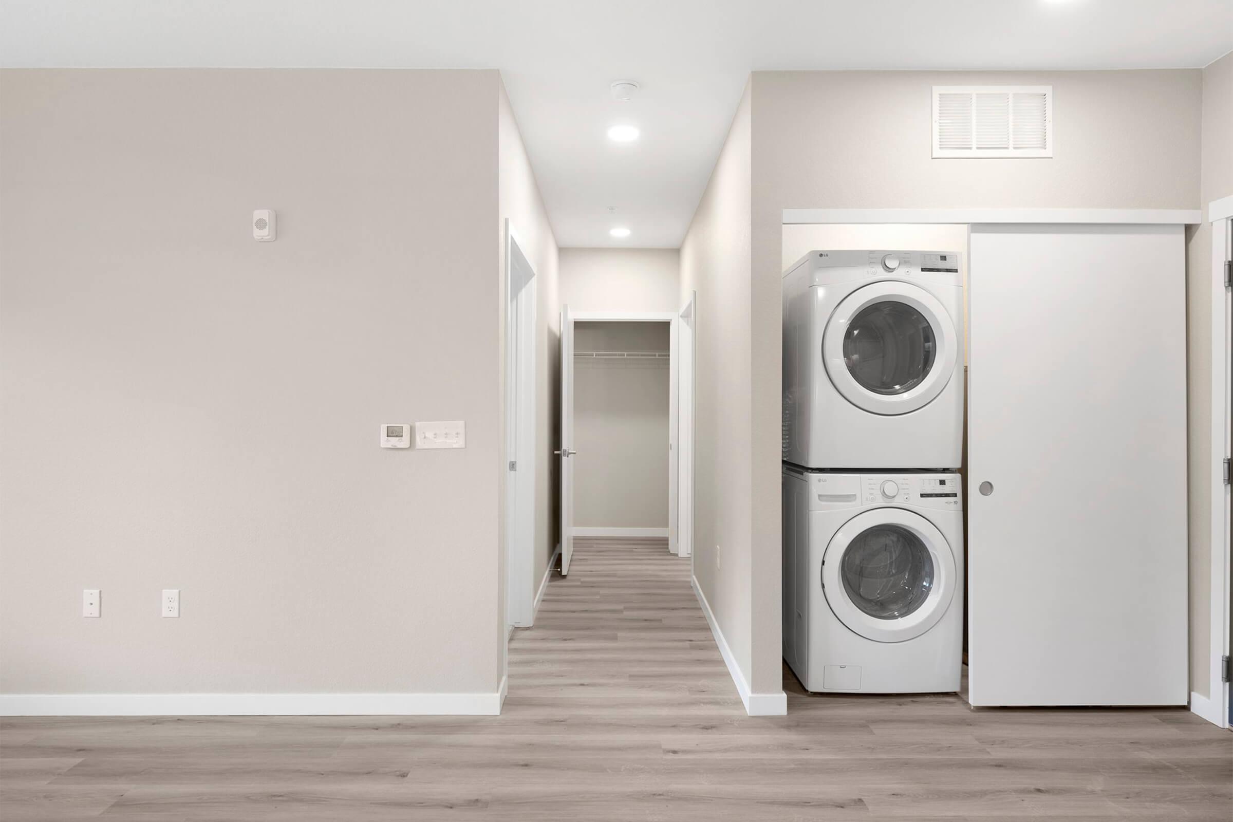 A modern laundry area featuring a stacked washer and dryer with a white finish. Light beige walls and a wooden floor create a clean, minimalist look. A hallway leads to a closet, enhancing the spacious feel of the room. Bright lighting adds to the inviting atmosphere.