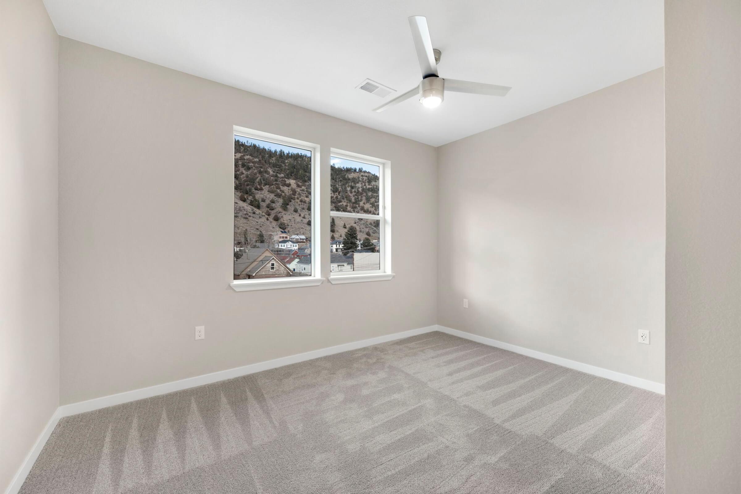 A well-lit, empty bedroom featuring light gray walls, large windows with a view of mountains, and a ceiling fan. The floor is covered with soft, gray carpeting, creating a serene and inviting atmosphere.