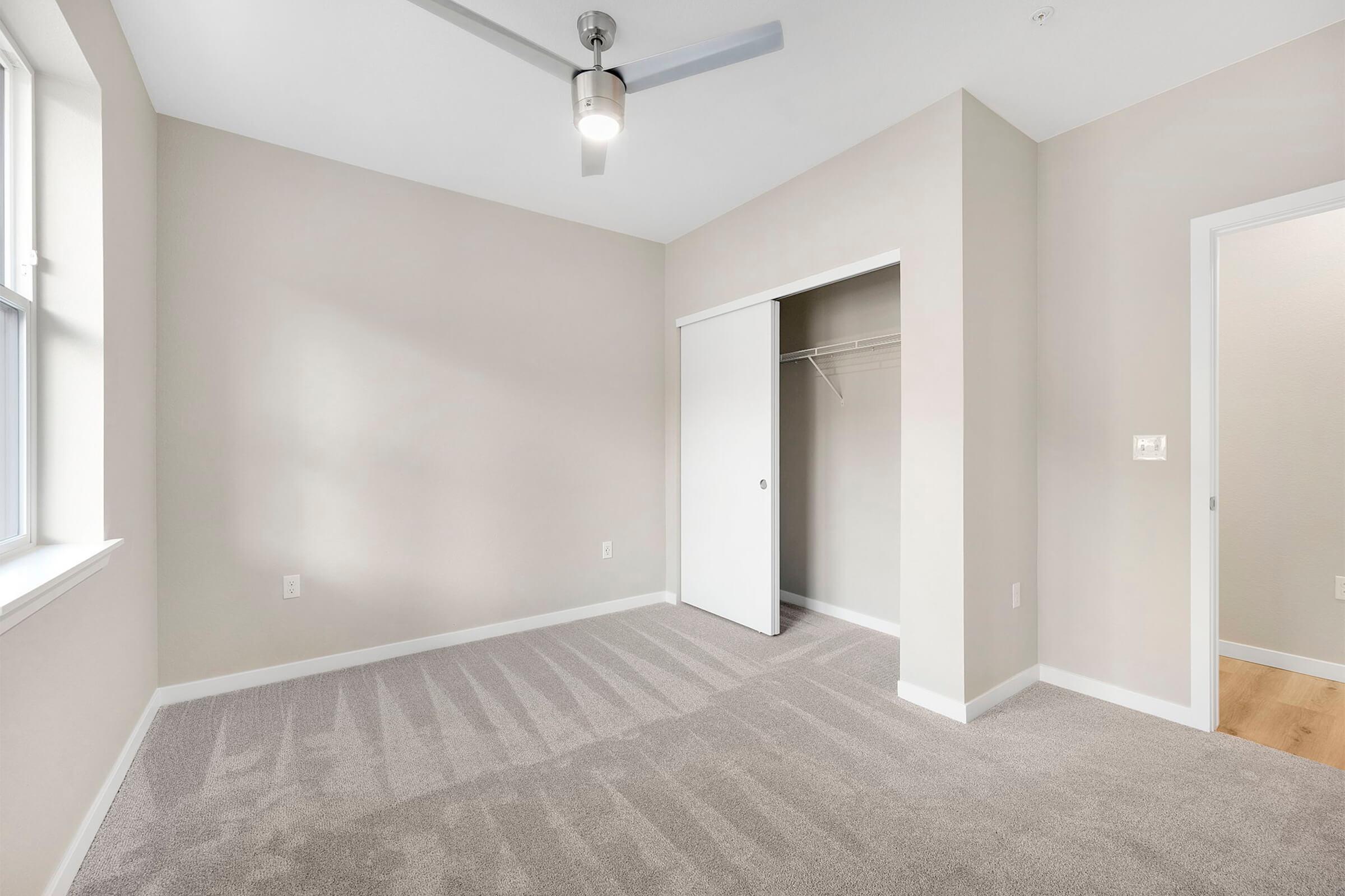 A well-lit, empty bedroom featuring neutral beige walls, a light carpet, and a ceiling fan. There is a closet with sliding doors on one side and a doorway leading to another room. Natural light streams in through a window, casting soft shadows on the carpeted floor.