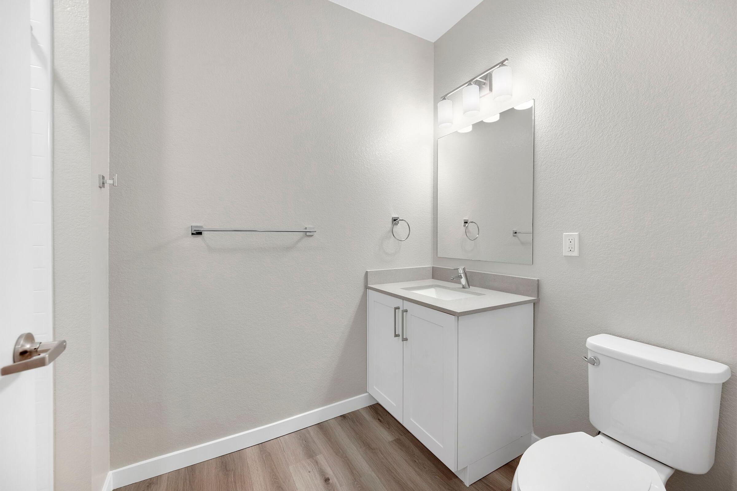 A modern bathroom featuring a white vanity with a rectangular sink and a large mirror above it. There is a toilet beside the vanity, a towel rack on the wall, and light-colored walls and flooring, creating a clean and spacious atmosphere. The door is slightly ajar, revealing more of the room.