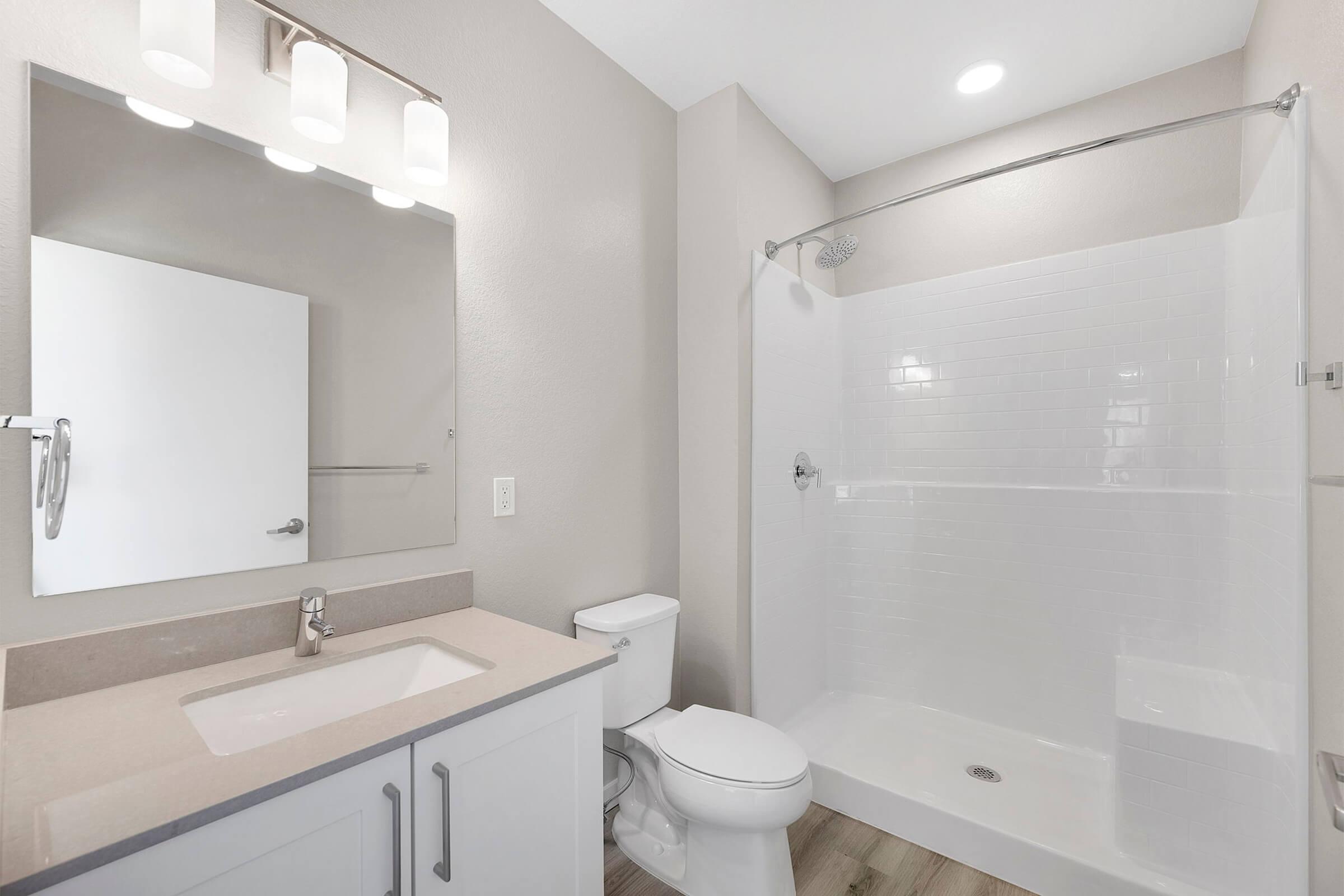 Modern bathroom featuring a white shower stall with glass doors, a sleek vanity with a countertop sink, a toilet, a large mirror above the vanity, and neutral-colored walls. Natural light illuminates the space, highlighting the clean and minimalist design.
