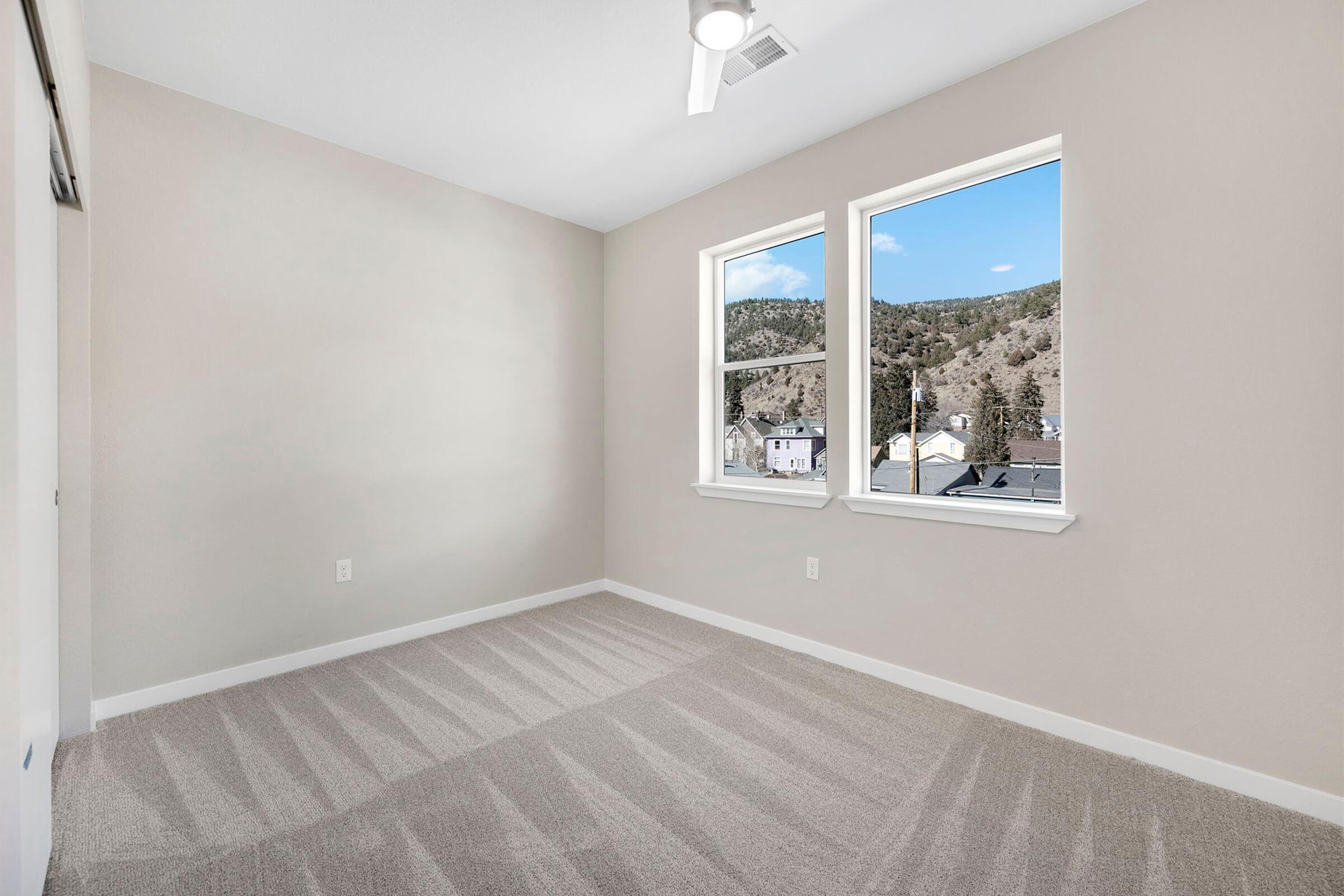 A well-lit, empty bedroom featuring two windows with mountain views. The walls are painted a light neutral color, and the floor is carpeted with a subtle stripe pattern. The ceiling has a modern ceiling fan.