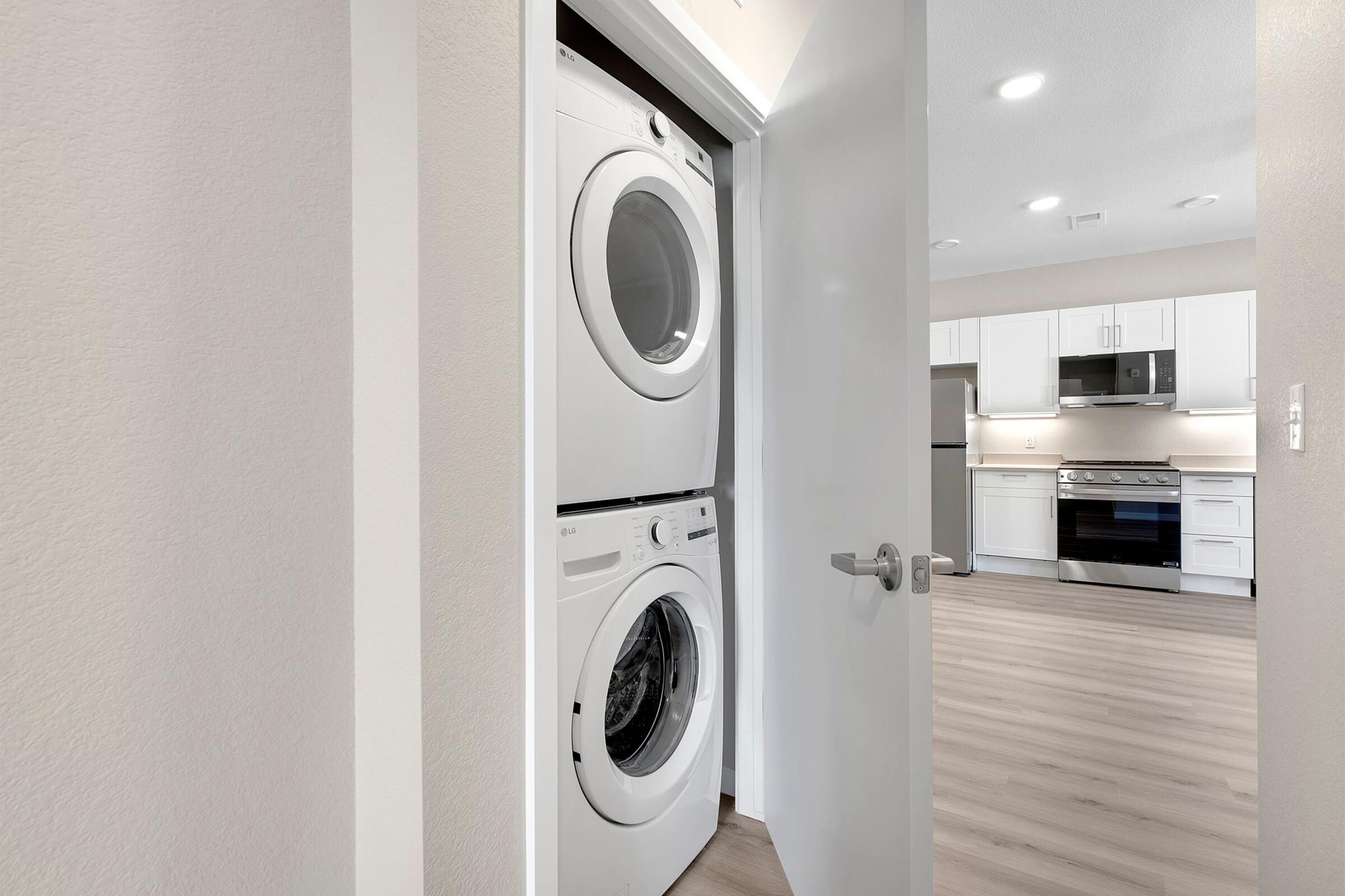 A stackable washer and dryer unit is visible in a white, open closet. The closet door is slightly ajar, revealing the laundry appliances. In the background, a modern kitchen with white cabinetry and stainless steel appliances can be seen, featuring laminate flooring.