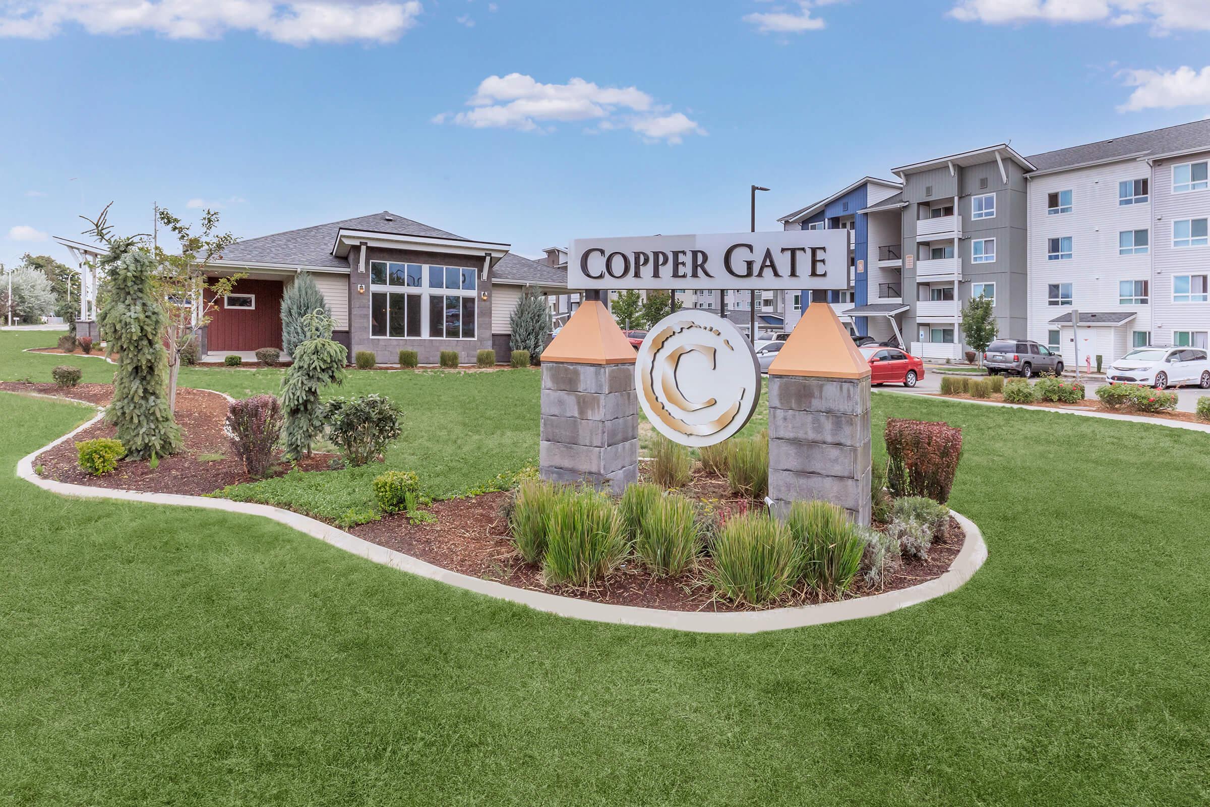 A well-maintained residential complex sign for Copper Gate, featuring a landscaped area with shrubs and grass. The sign is set between two decorative pillars with orange tops. In the background, there are modern apartment buildings and a clear blue sky.