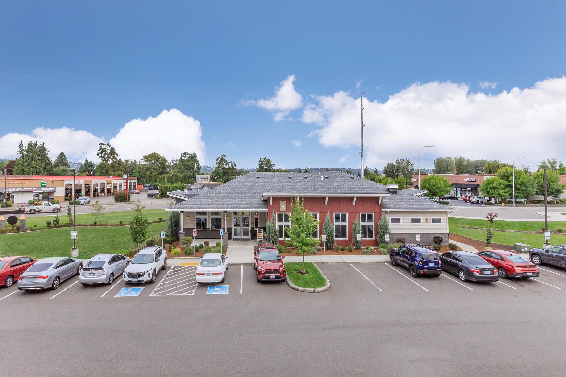 A modern single-story building with a gray roof, surrounded by landscaped areas and parking spaces. Several cars are parked in front, including accessible parking. In the background, additional buildings and green trees are visible under a clear blue sky with some clouds.