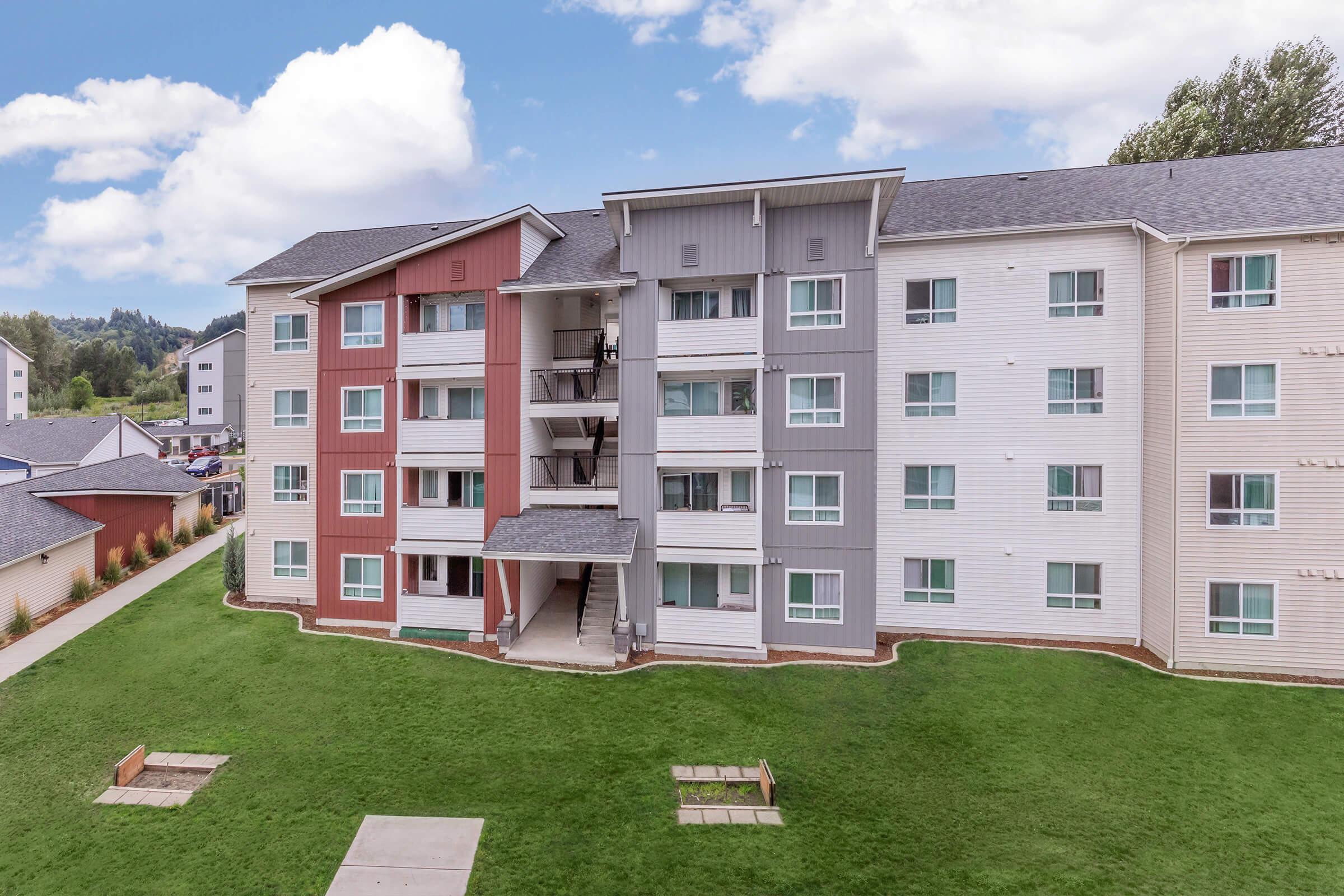 A modern multi-story apartment building with a mix of red, gray, and white exterior panels. The building features balconies, large windows, and a well-maintained grassy area in front, accompanied by a pathway. The sky is partly cloudy, and the surrounding landscape includes additional residential buildings.