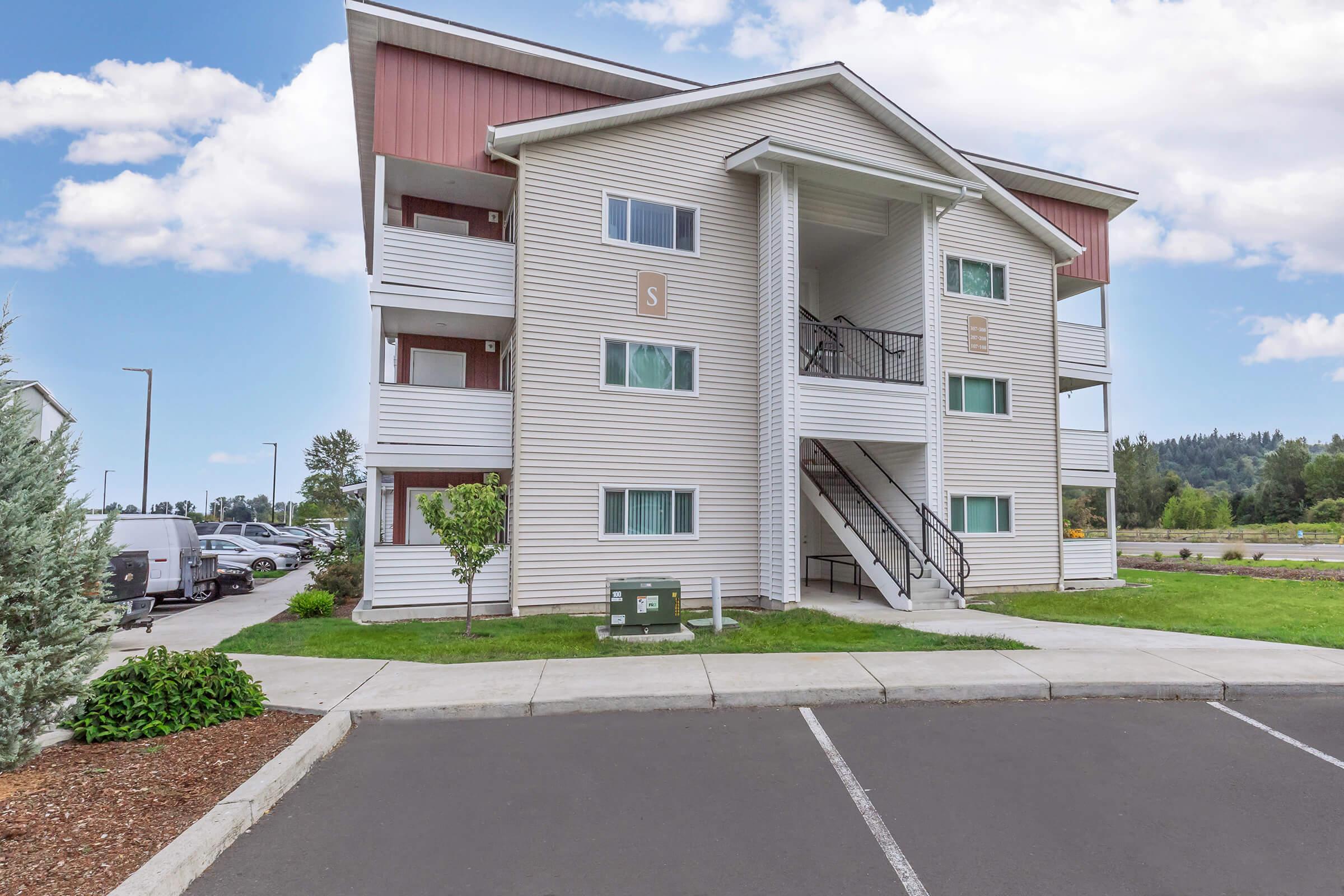 A three-story residential building with a modern design, featuring a light-colored exterior and a red accent on the roof. The building has balconies on the upper floors, a central staircase, and nearby grassy areas with trees. A parking lot is visible in front, with several parked vehicles.