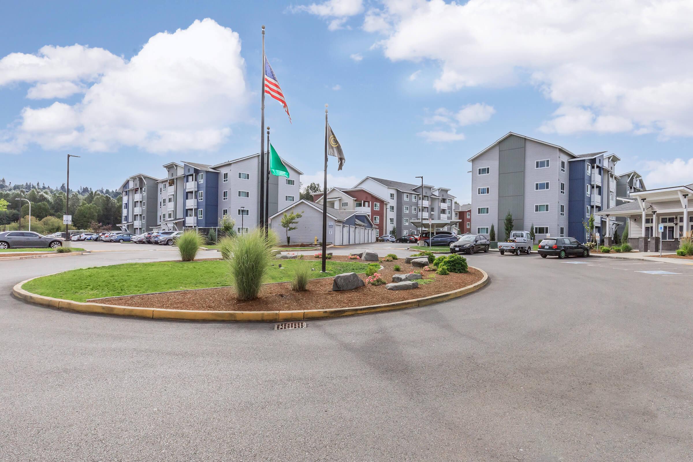 A circular driveway with landscaped roundabout featuring flags. Modern apartment buildings are visible in the background, surrounded by greenery and parked cars. The sky is bright with scattered clouds, creating a welcoming atmosphere.