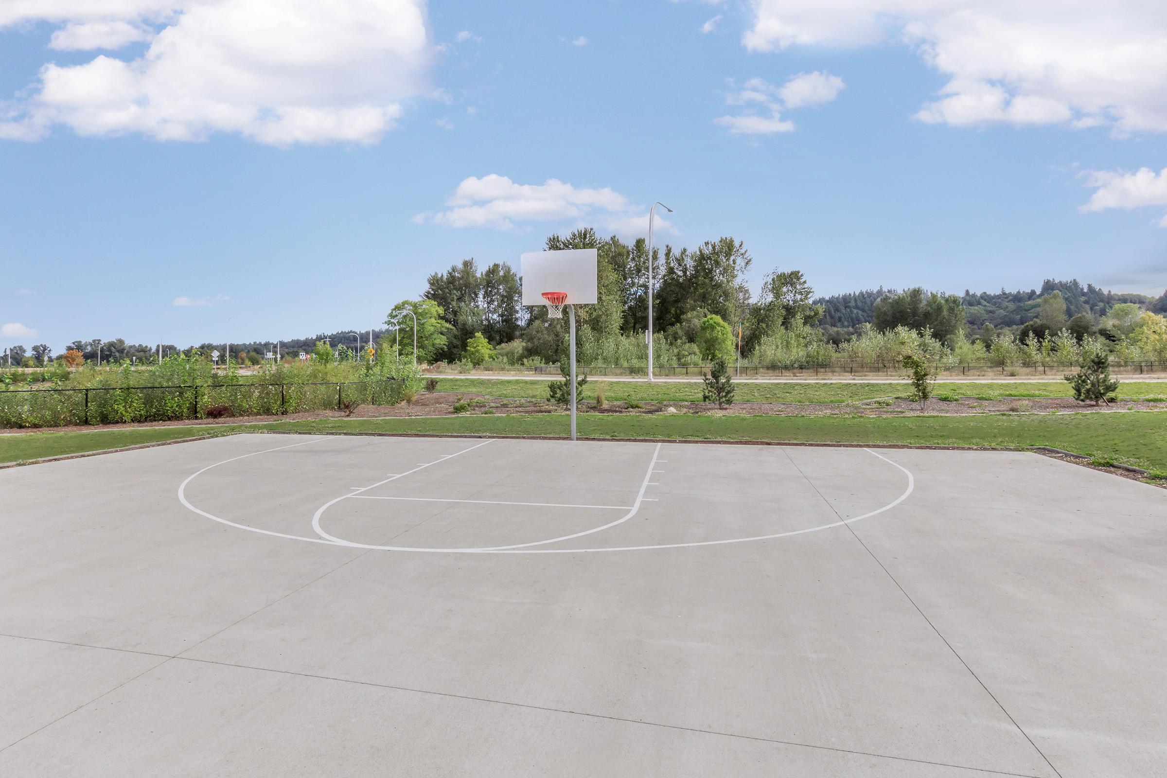 A basketball court featuring a single hoop and a smooth concrete surface, surrounded by greenery and trees. The sky is clear with a few clouds, creating a bright and open atmosphere. The court is empty, emphasizing its inviting space for players.