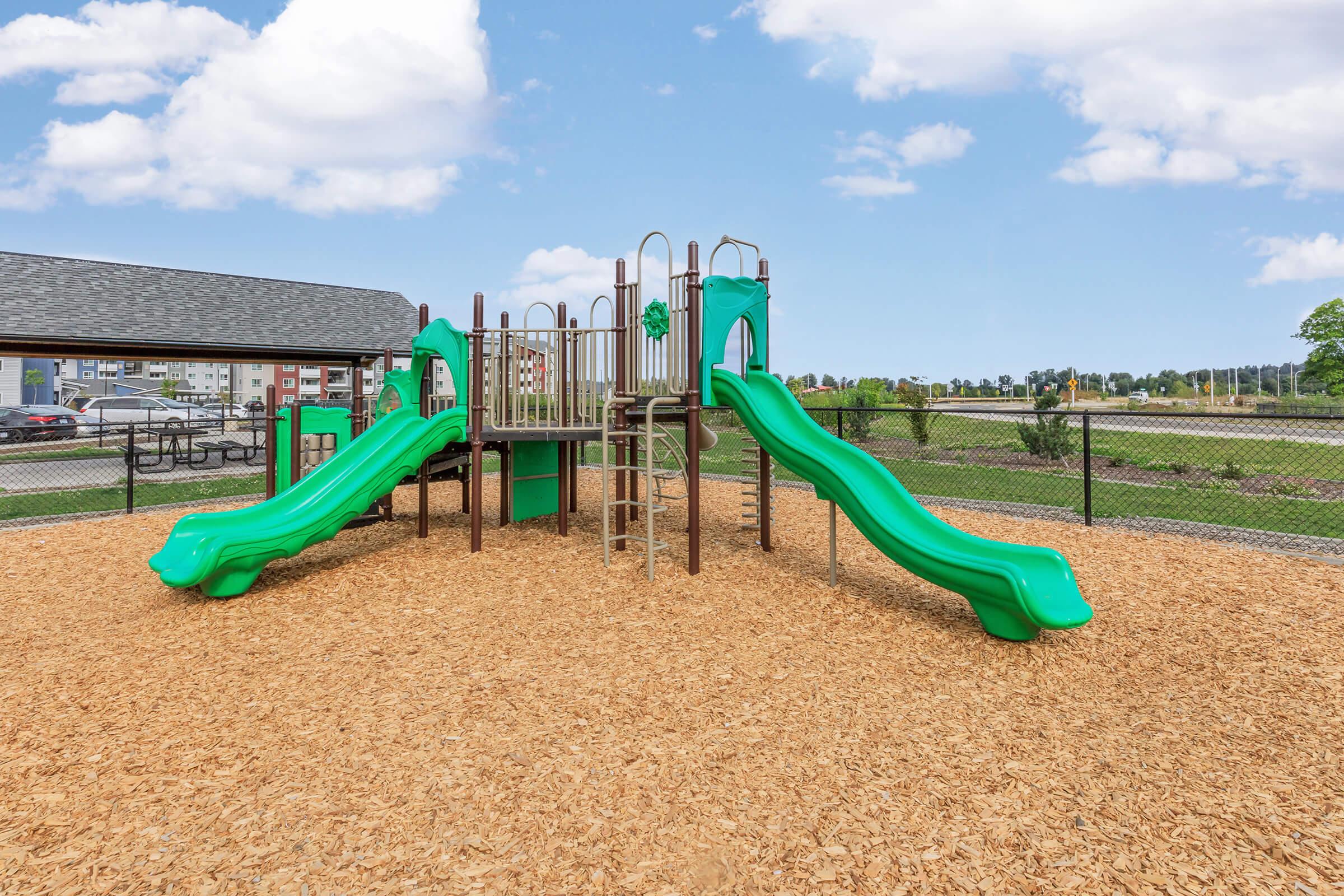 A playground featuring two green slides, a climbing structure, and safety features, surrounded by wood chips. In the background, there are buildings and a grassy area, with a blue sky and scattered clouds above.