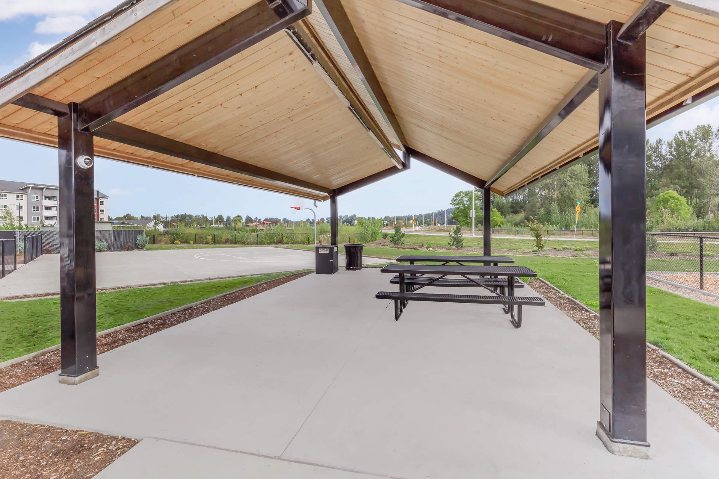 A covered outdoor pavilion with a wooden roof and black support beams, featuring picnic tables beneath. In the background, there's a grassy area and a basketball court, with trees and buildings visible in the distance. The scene is bright and open, ideal for gatherings or recreational activities.