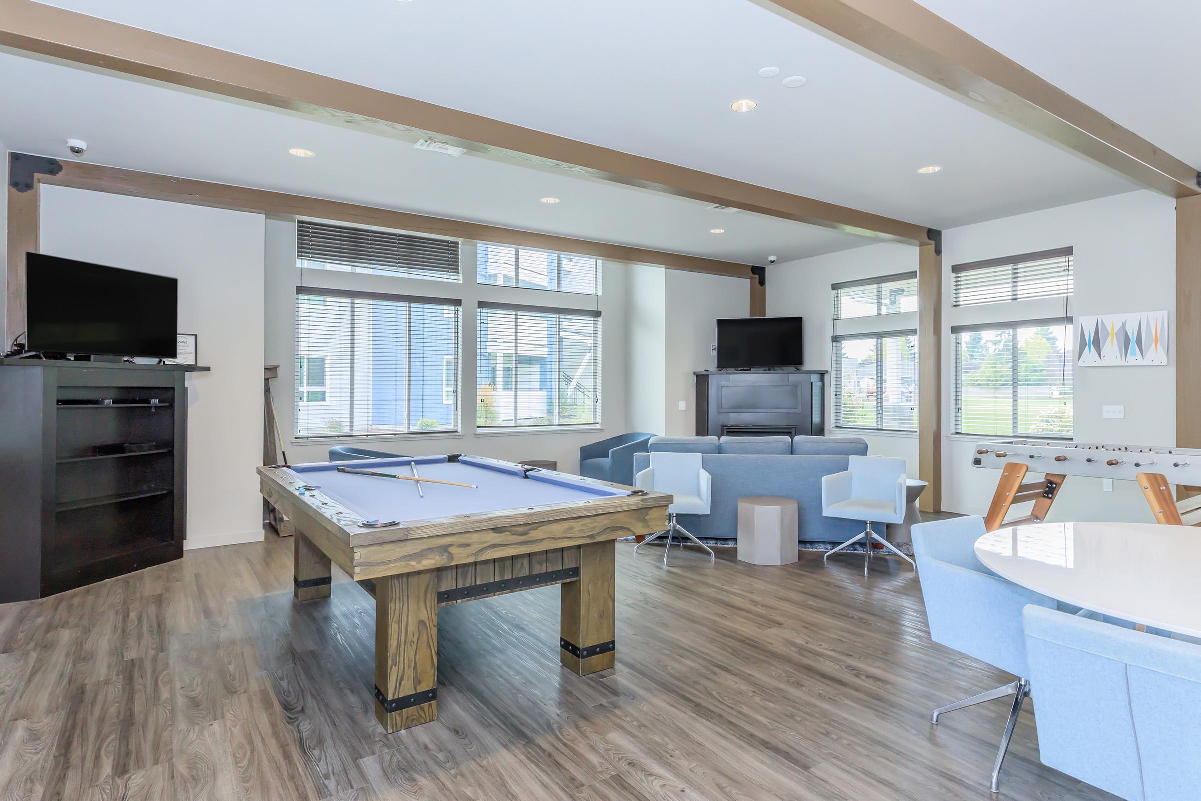 A modern communal lounge area featuring a pool table, comfortable seating with blue chairs and gray sofas, a round dining table, large windows providing natural light, and two flat-screen TVs. The floor is covered with light-colored wood laminate, creating a spacious and inviting atmosphere.
