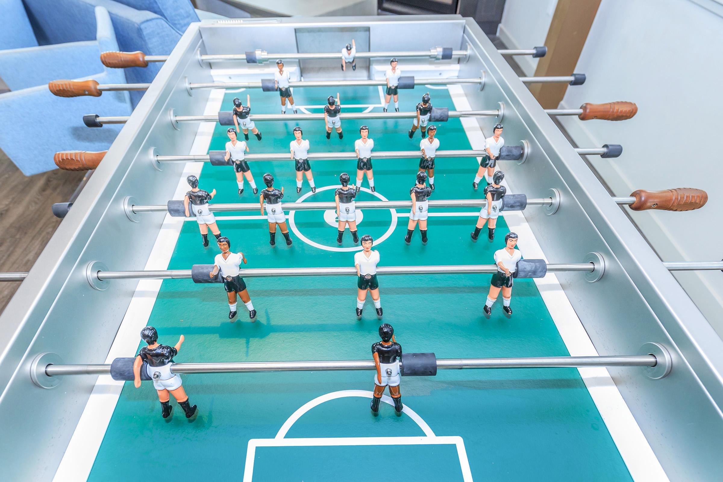 A foosball table featuring multiple players arranged in teams, with figures dressed in black and white uniforms. The playing field is green, marked with a soccer pitch layout. The rods and handles for player manipulation are visible, highlighting a classic game setup.
