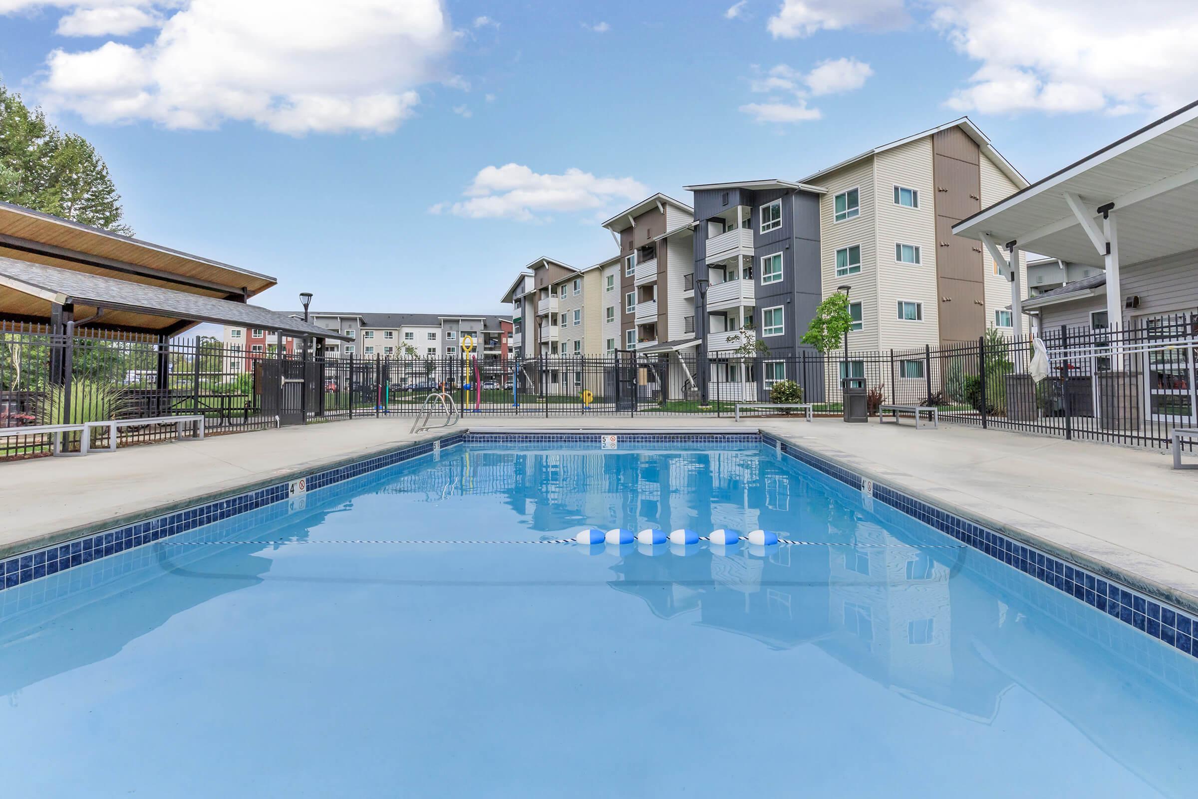 A clear swimming pool with a blue tiled edge, surrounded by a fenced area. In the background, there are modern apartment buildings and a shaded pergola. The sky is partly cloudy, contributing to a bright and inviting atmosphere.