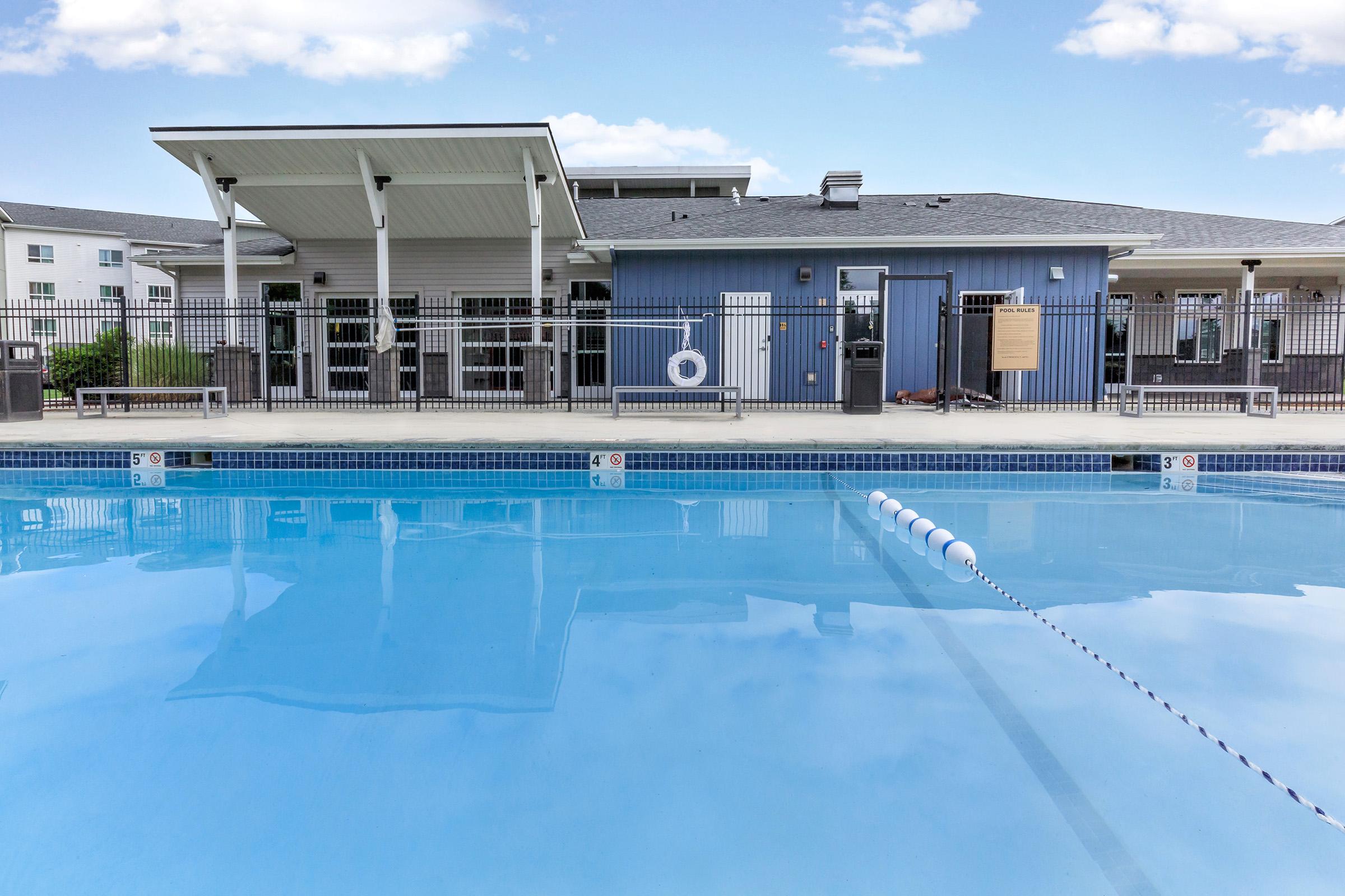 A clear swimming pool with a floating lane marker, surrounded by a fenced area. In the background, there is a modern building featuring large windows and a shaded patio area. The sky is bright with a few clouds.