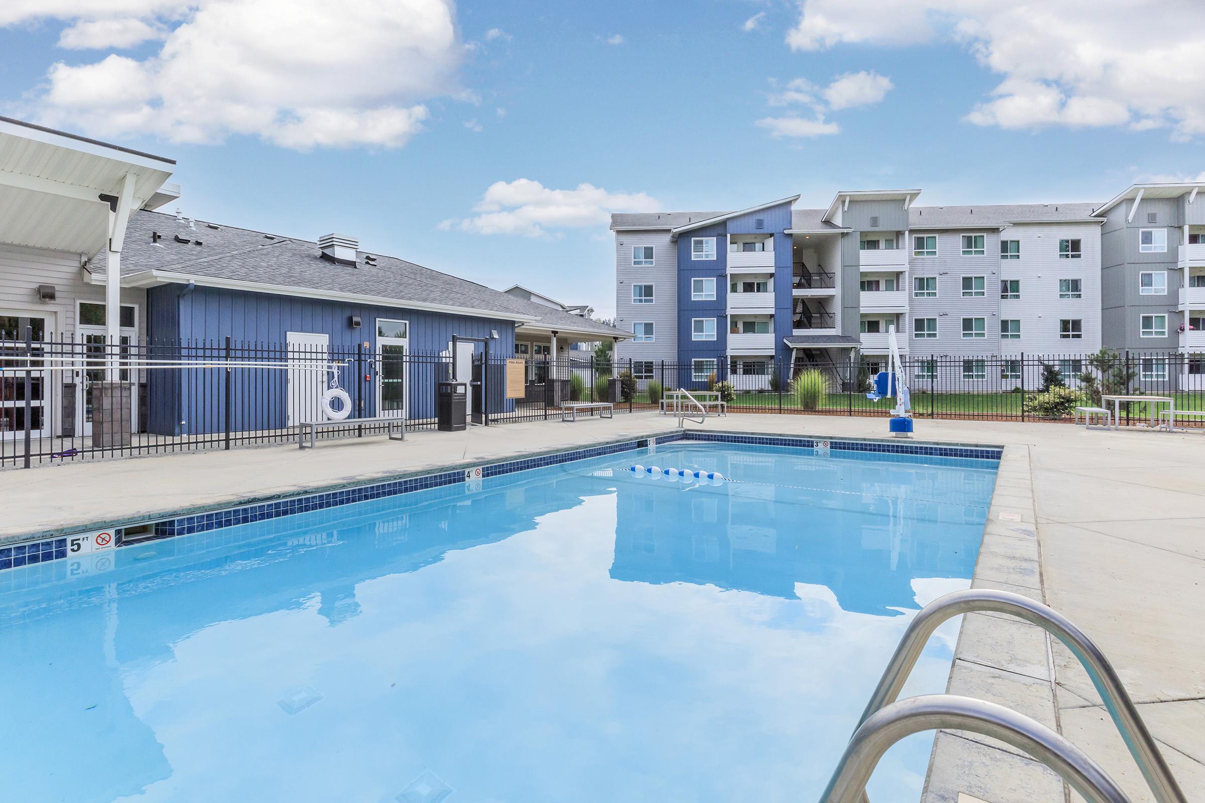 Swimming pool with clear blue water, surrounded by a concrete deck and lounge area. Nearby buildings with modern architecture are visible, featuring multiple stories and large windows. A sunny sky with scattered clouds completes the scene, creating a relaxing atmosphere.