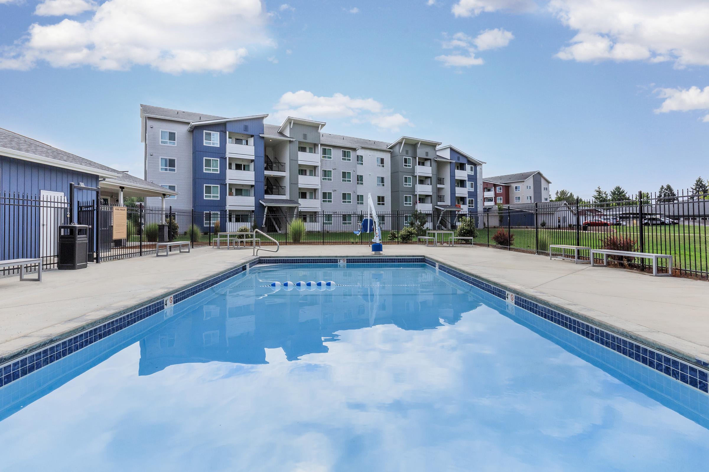 A clear blue swimming pool surrounded by a well-maintained deck, with lounge chairs and an umbrella. In the background, three multi-story residential buildings with a mix of blue, gray, and white colors, fenced area, and green lawn. Bright, sunny sky with scattered clouds.