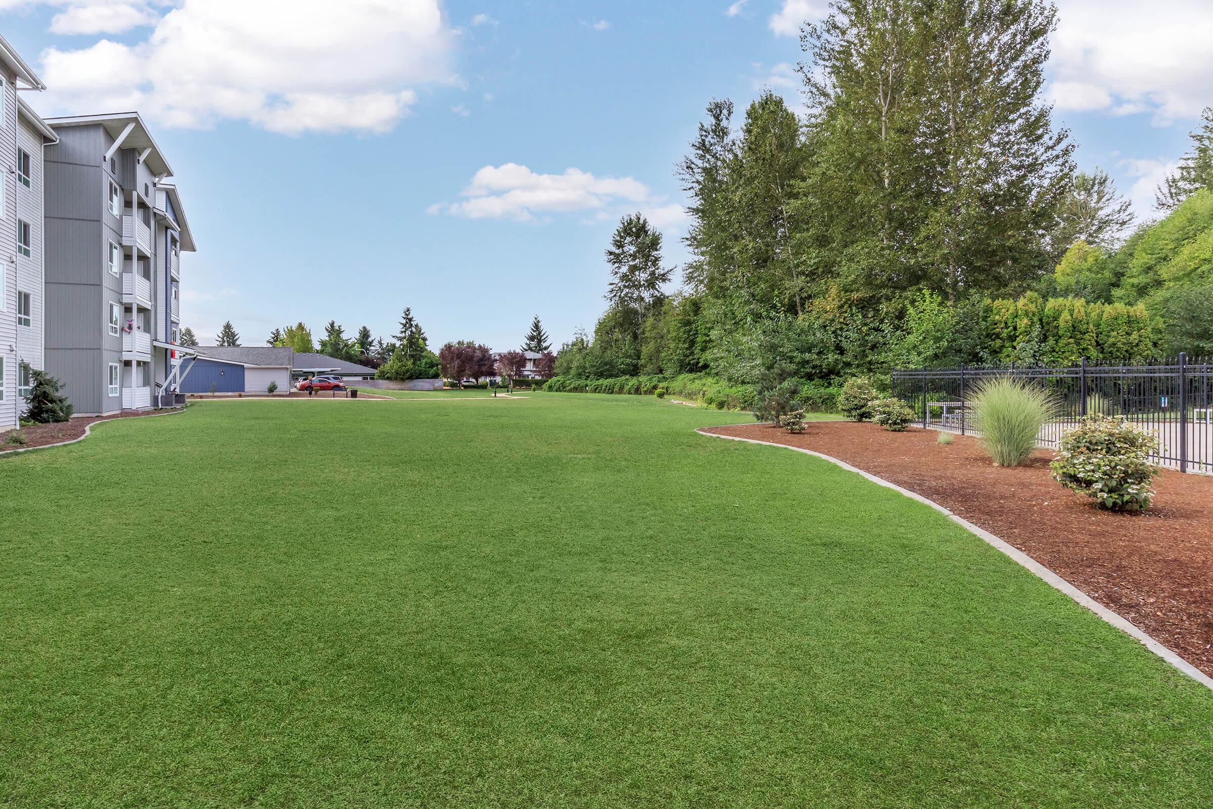 A well-maintained green lawn next to an apartment building, bordered by trees and shrubs. In the distance, there are benches and a fenced area, with a clear blue sky above and a few fluffy clouds. The setting appears peaceful and inviting, ideal for outdoor activities or relaxation.