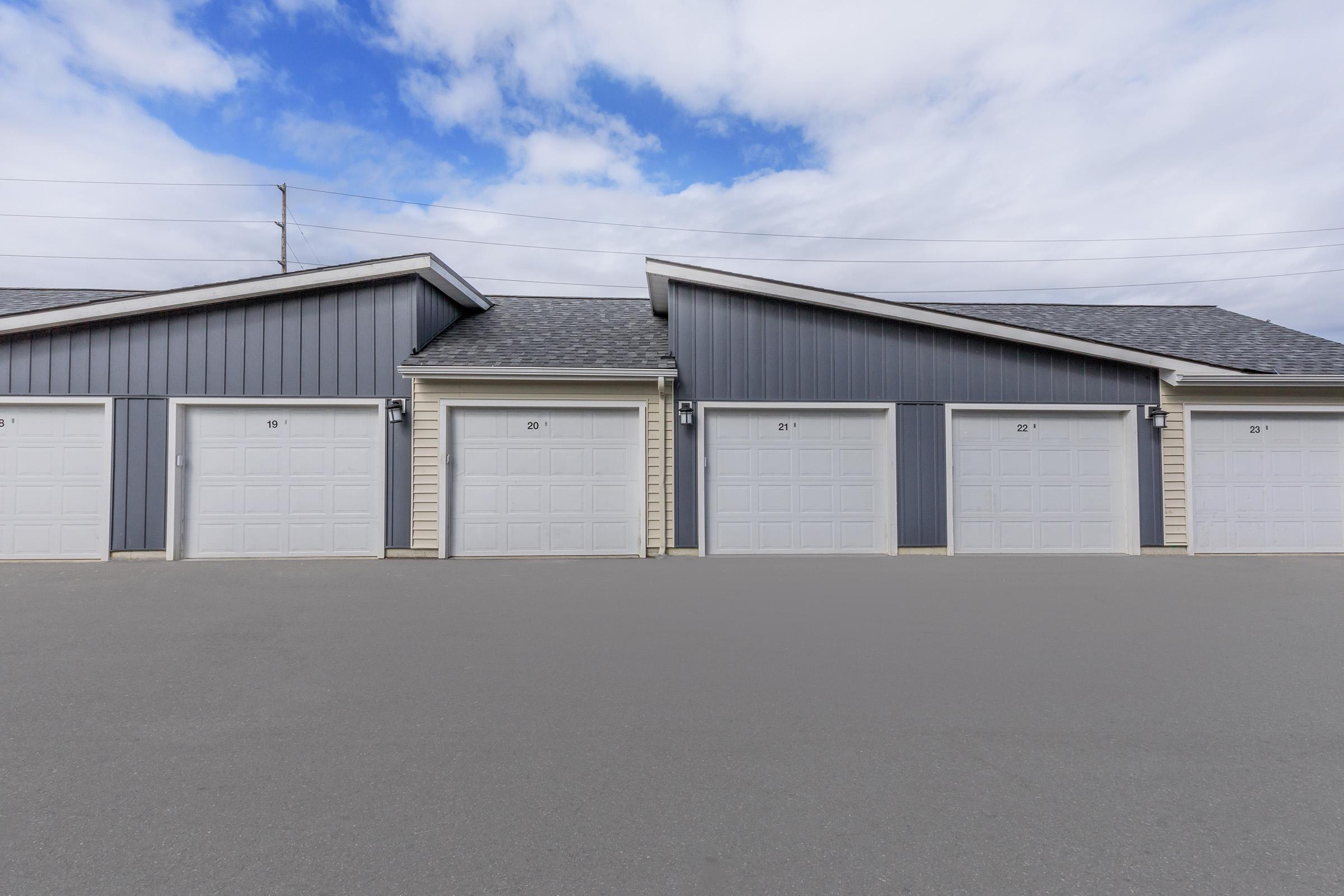 Four gray garage doors lined up in a row, with the numbers 19, 20, 21, and 22 visible on each door. The building has a simple, modern design with a sloped roof, and the sky above is partially cloudy. The pavement in front is smooth and unmarked.